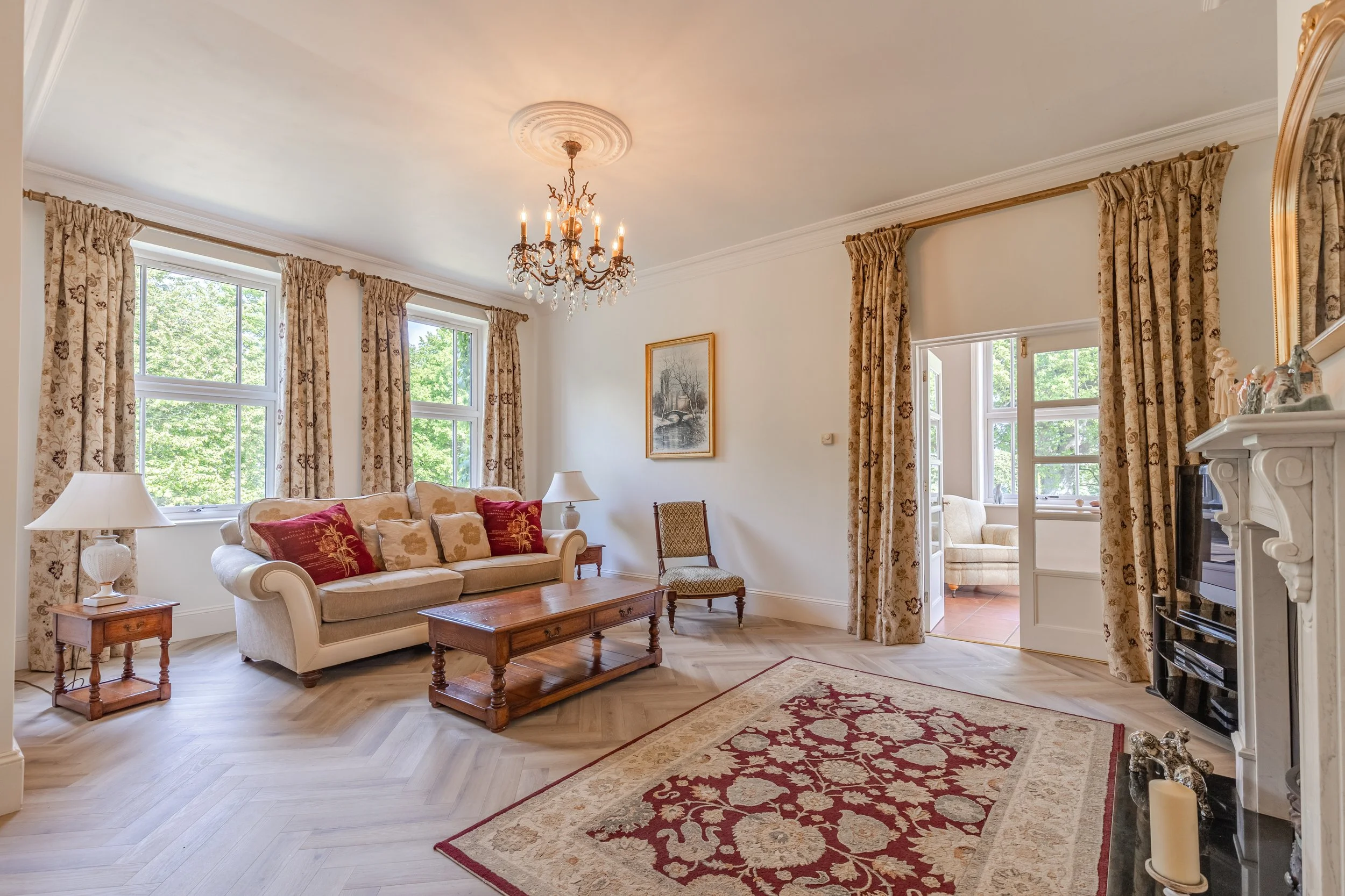 A living room with a white sofa decorated with red and beige pillows, a wooden coffee table, a side table with a white lamp, framed artwork on the wall, large windows with floral curtains, a fireplace with decorative objects, and a chandelier hanging