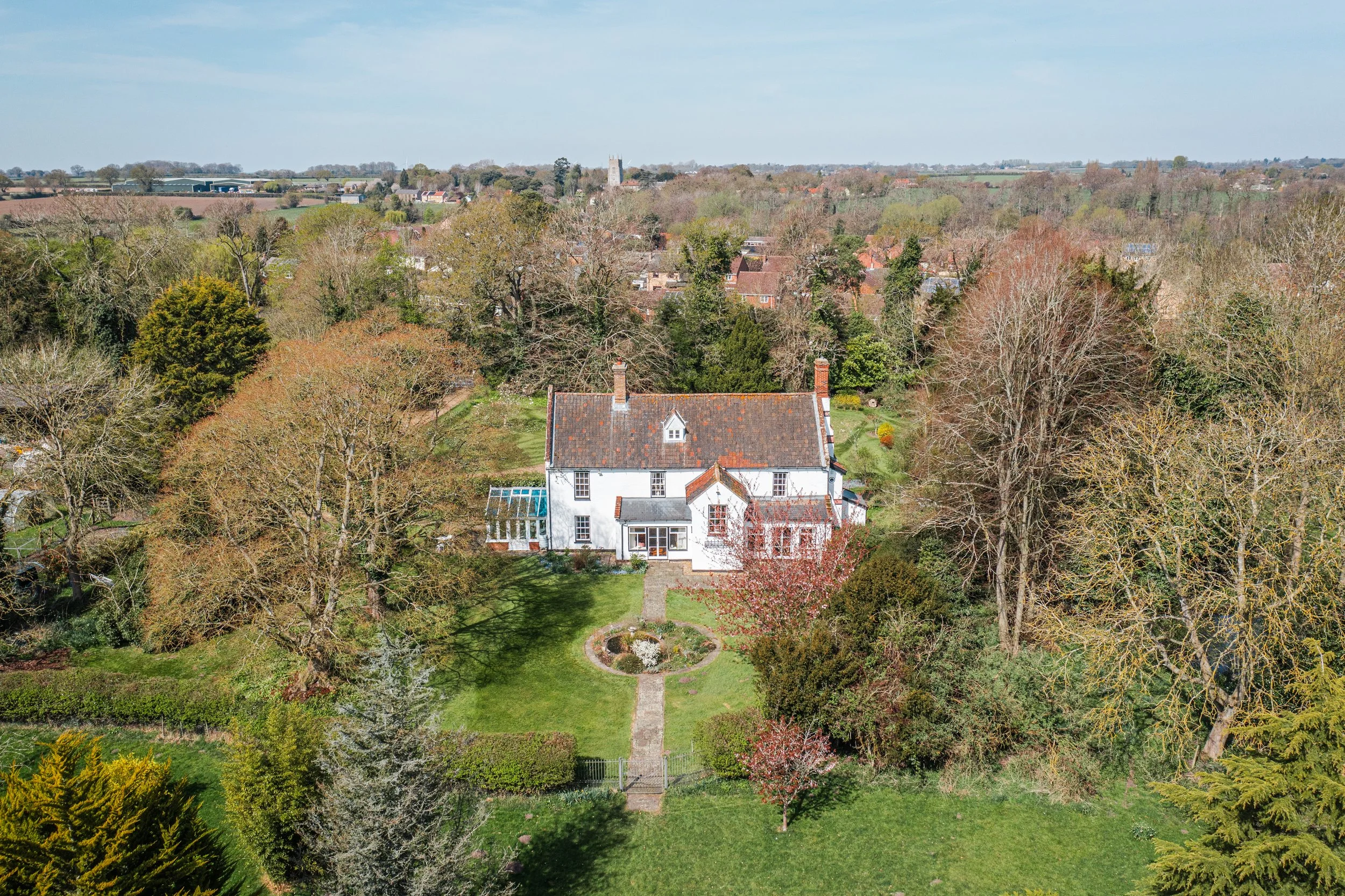 Aerial view of a large white house with a garden, surrounded by trees with some leaves changing color, with a village and rolling green hills in the background under a blue sky.