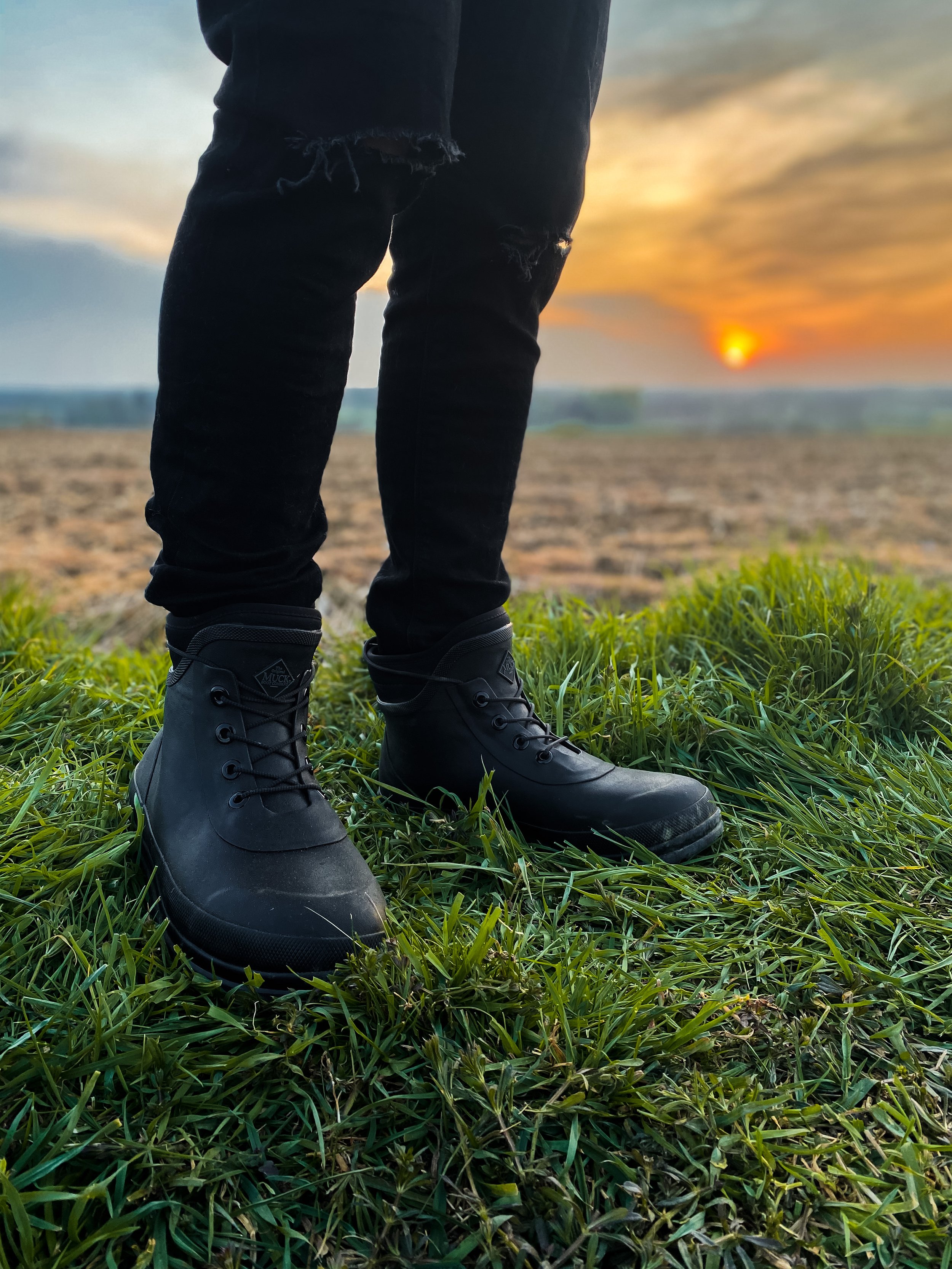 Close-up of black combat boots worn by person standing on green grass with a beach and sunset in the background, wearing ripped black jeans.