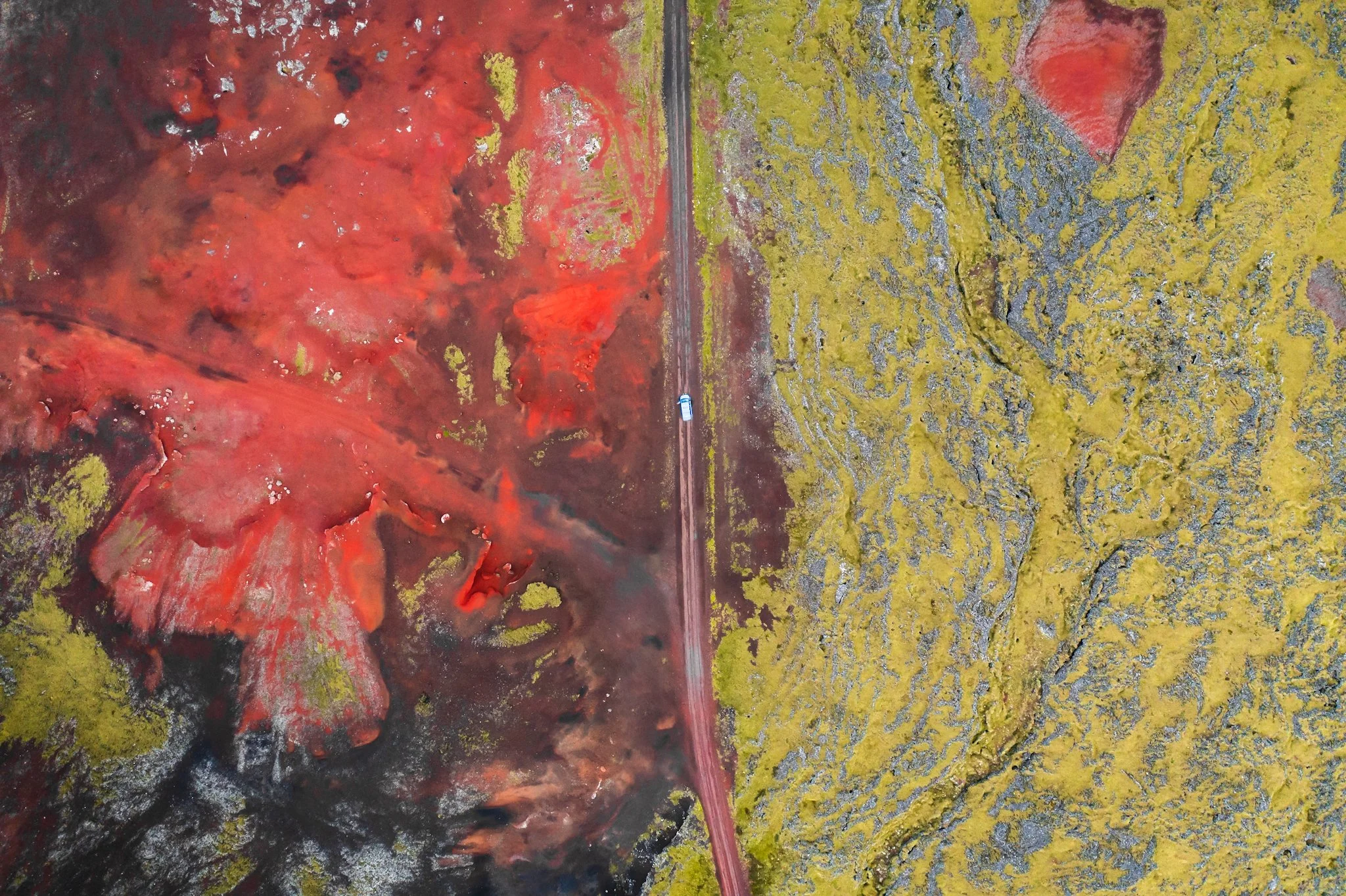 Aerial view of an abstract volcano in Iceland. The left side features a fiery red and black swirl with streaks of white, while the right side depicts a yellow and gray textured pattern with a distinct red triangle near the top right corner.