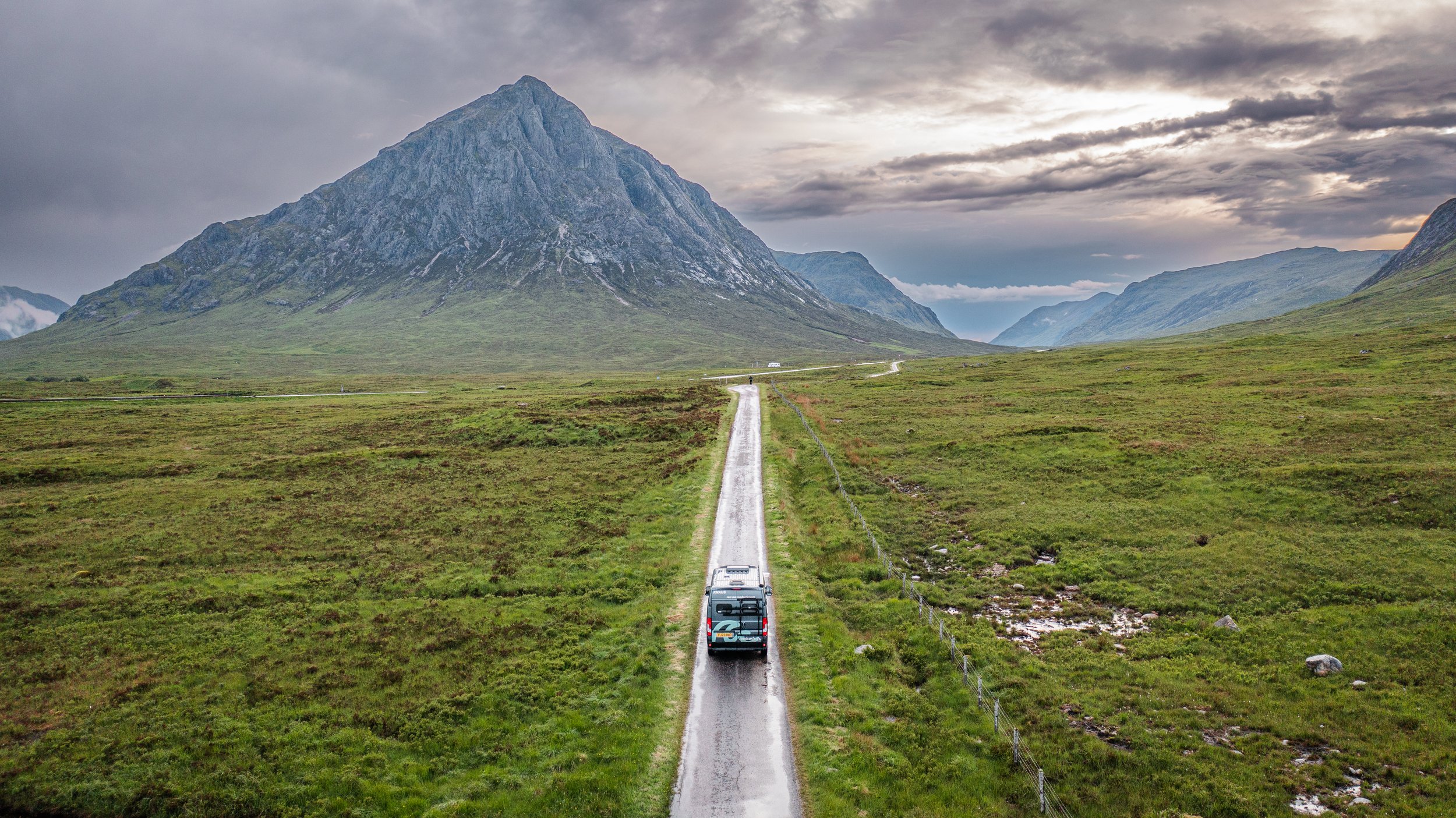 A vehicle driving on a narrow road through lush green plains, with a mountain in the background and a cloudy sky overhead.