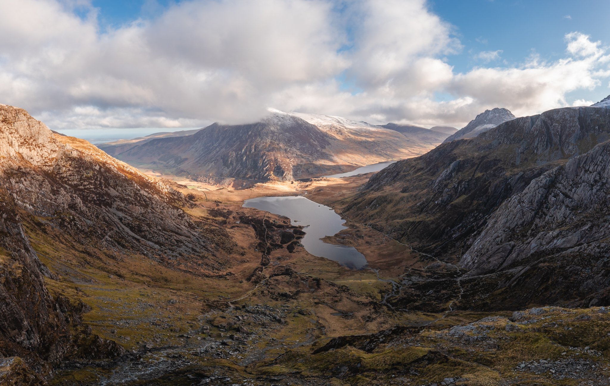 Mountain landscape with a lake, cloudy sky, and snow-capped peaks.