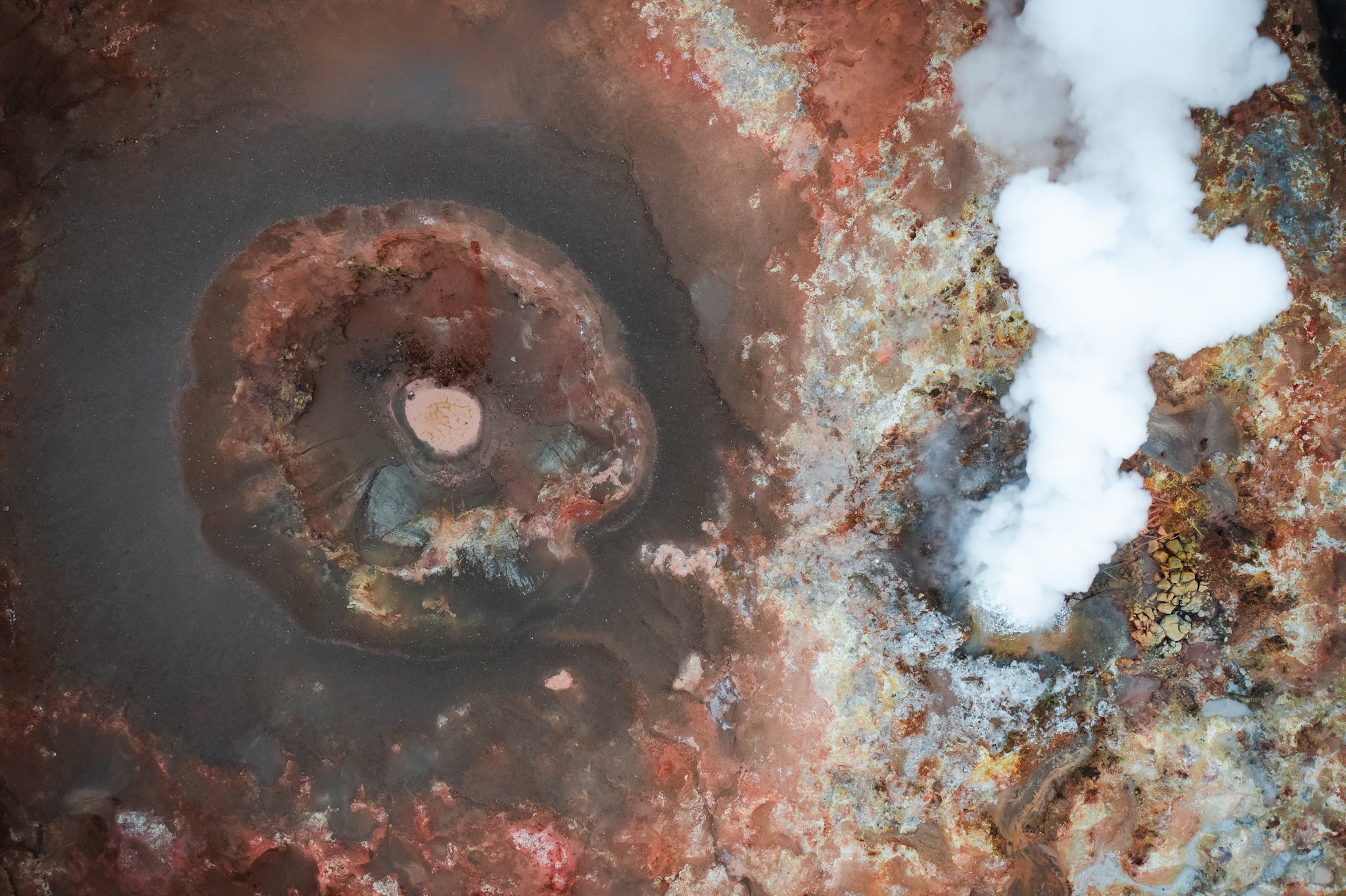 A geothermal hot spring or fumarole emitting white steam in a volcanic landscape with colorful, mineral-rich rocks.