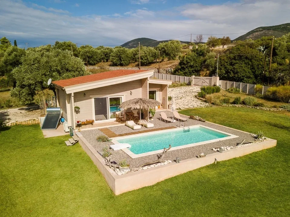 A backyard with a swimming pool, lounge chairs, and outdoor umbrella in front of a modern house, with green grass, trees, and hills in the background.