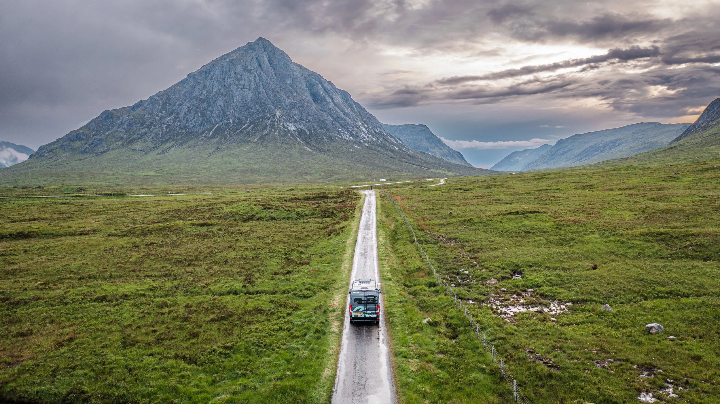 A narrow road with a vehicle driving on it, stretching through green fields towards a large mountain with cloudy skies overhead.
