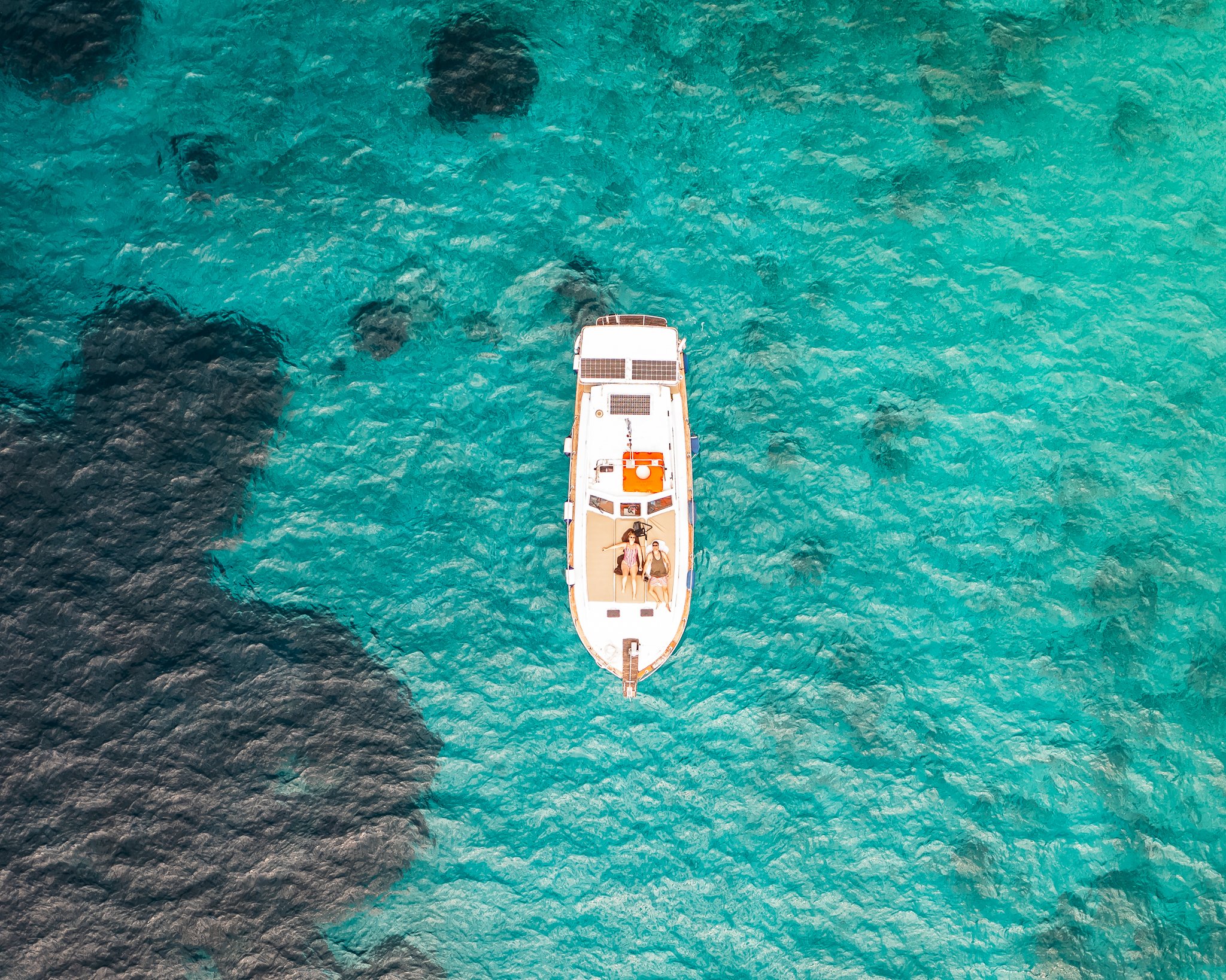 An aerial view of a boat with several people on board, sailing through clear turquoise water with visible rocks beneath.