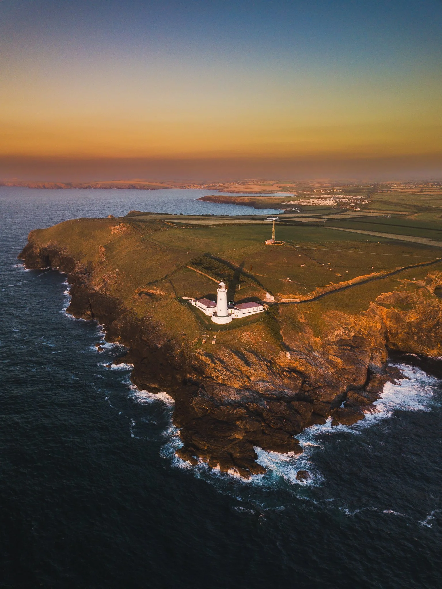 A lighthouse on a rocky coastline during sunset, with a vast ocean on one side and green fields with farmland on the other.