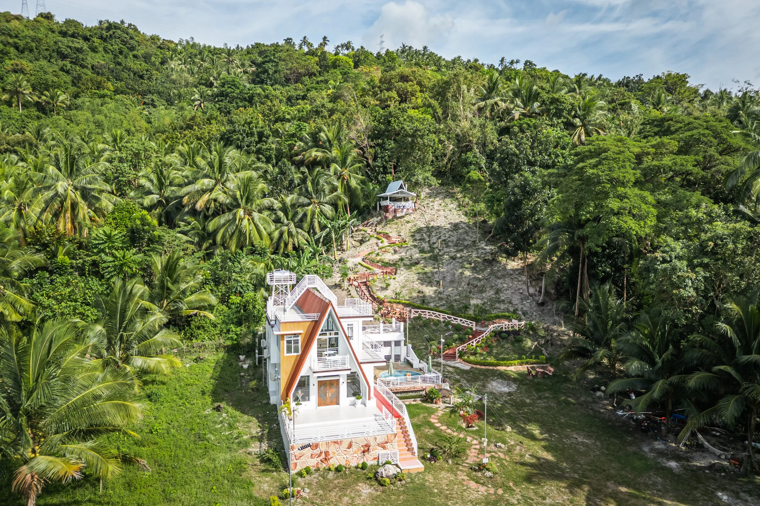 A modern multi-story house with white exterior walls, large windows, and a rooftop terrace, situated on a hillside surrounded by lush green tropical trees and vegetation.