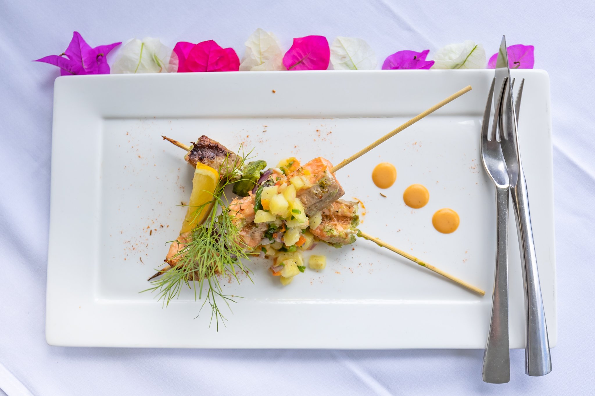 A white rectangular plate contains a small serving of fish topped with chopped vegetables, garnished with fresh dill, and decorated with three drops of orange sauce. There are broken pieces of fish on a skewer. Pink and white flowers are arranged alo