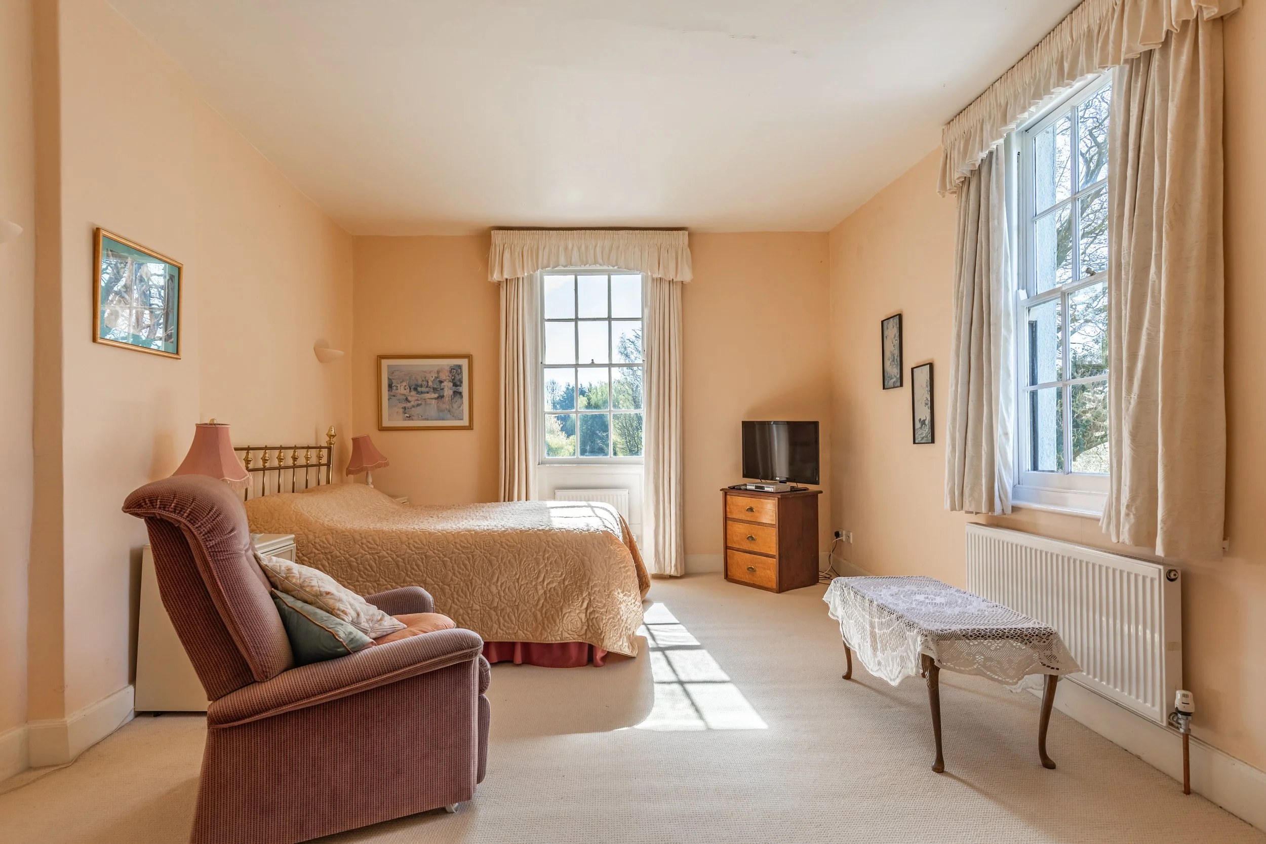 Bright bedroom with a large window, cream-colored walls, a bed with a peach quilt, a pink armchair, a small dresser with a TV, framed paintings, and white curtains.