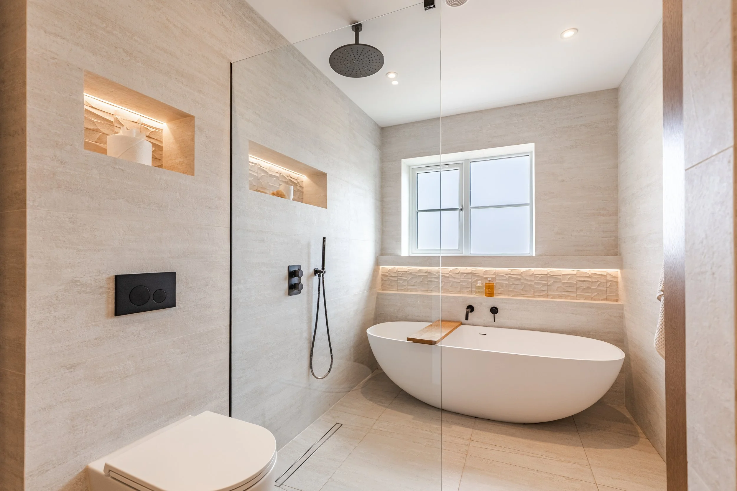  Modern bathroom with a bathtub, shower area with a rainfall showerhead, built-in shelves with decor, frosted window, and neutral-toned tiles.