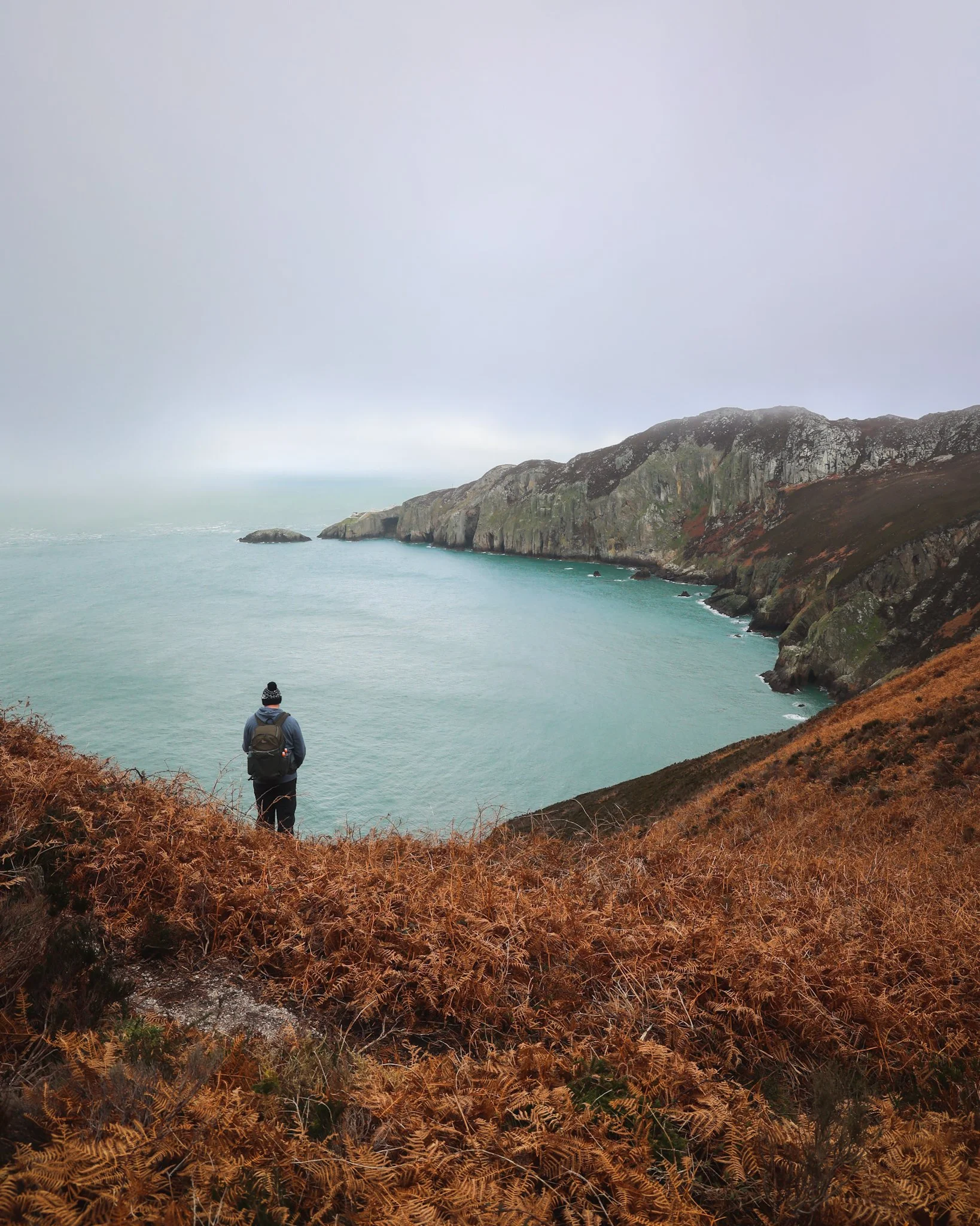 A person with a backpack and hat looking at the ocean from a cliff. The coastline has large rocks and a steep hillside covered in brown foliage, with fog over the water and hills in the distance.