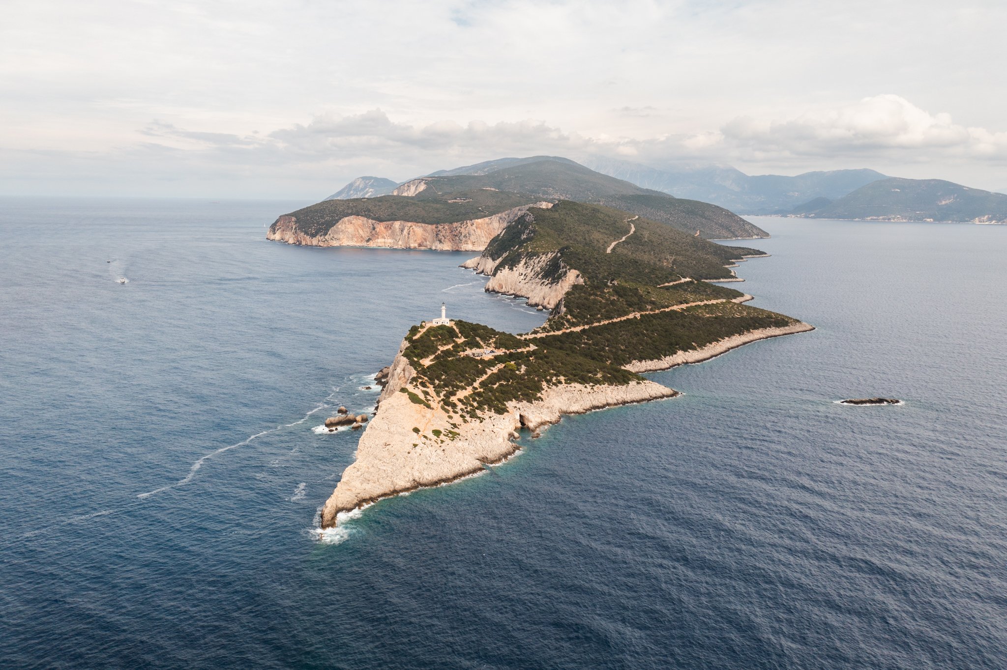 Aerial view of a coastal landscape with green-covered cliffs extending into the ocean, a lighthouse on a promontory, and boats on calm water under a cloudy sky.