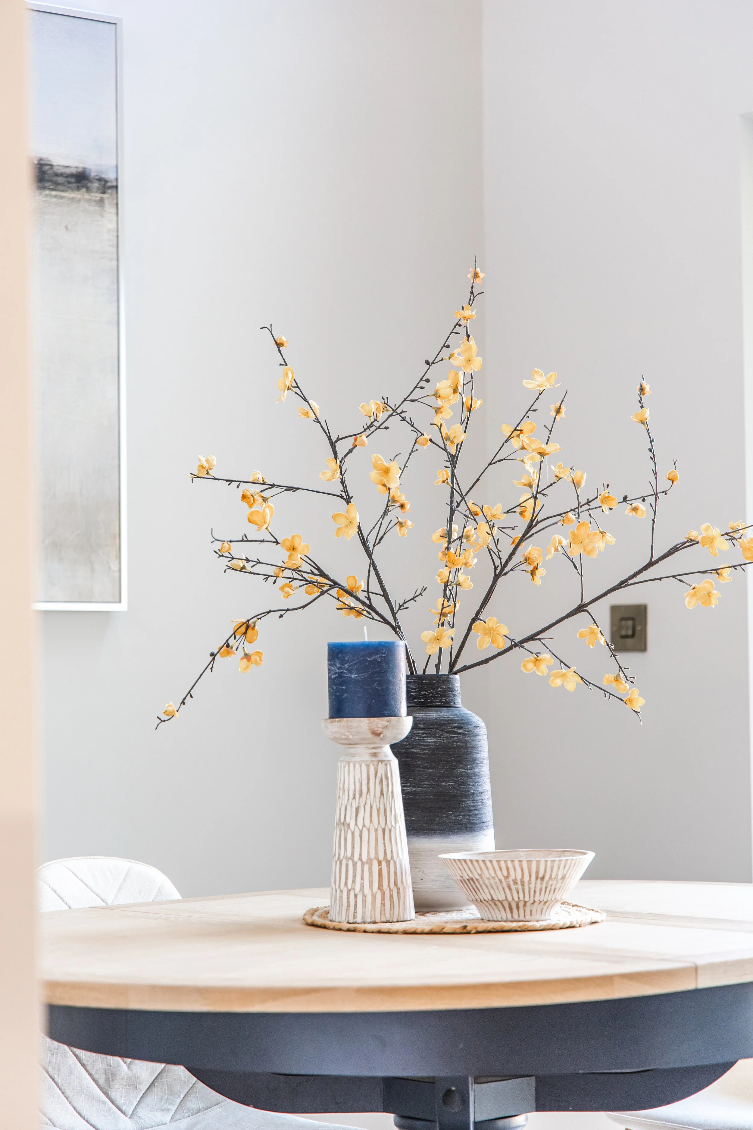 A round table with a wooden top holds three decorative vases of different shapes and sizes, with a branch of yellow flowers in one of the vases. The table is in a bright room with white walls and a mirror or window on the left side.