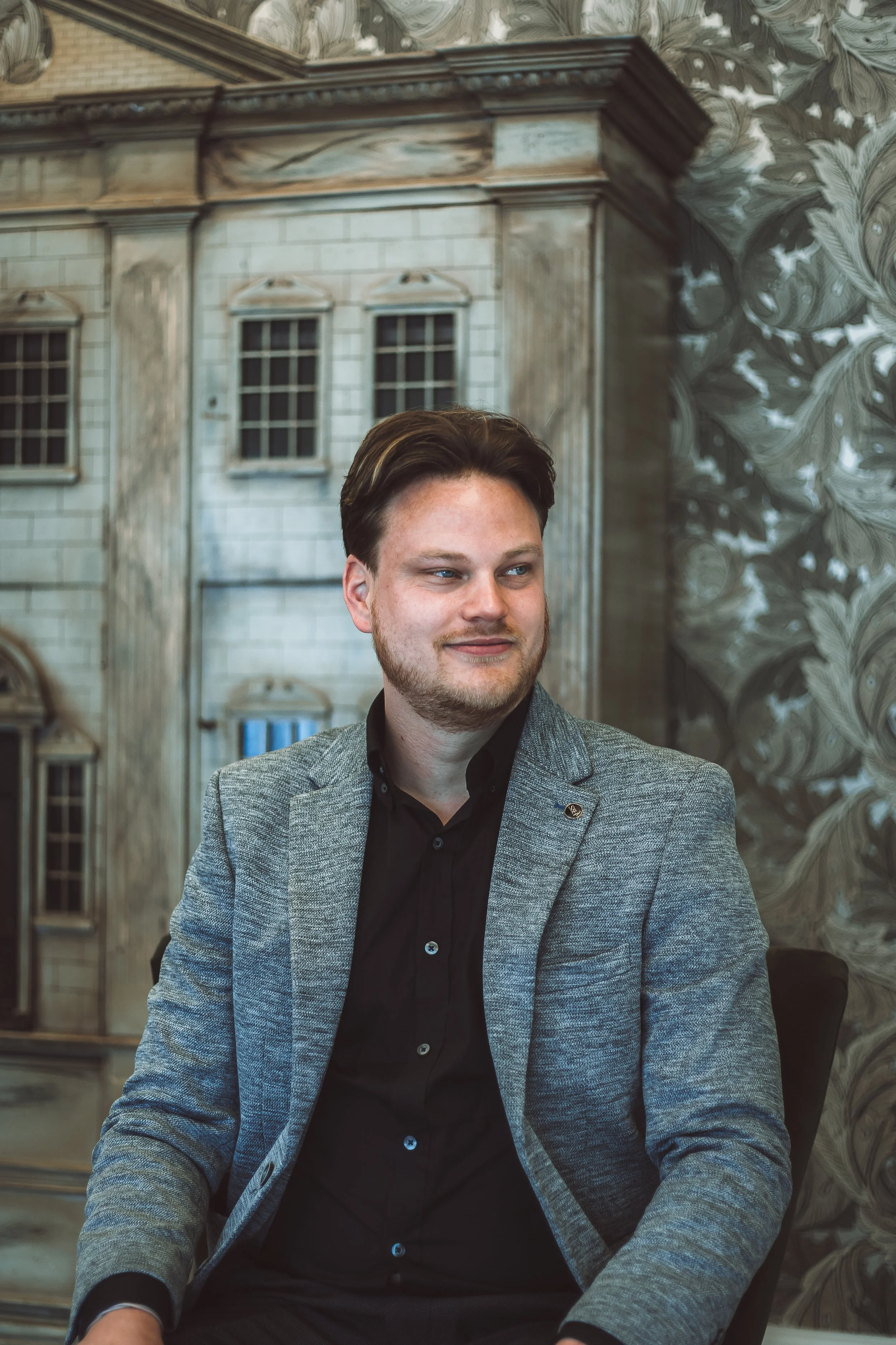 A young man with brown hair and a beard sitting indoors in front of a detailed decorative wall and an ornate building facade background, wearing a gray blazer and black shirt.