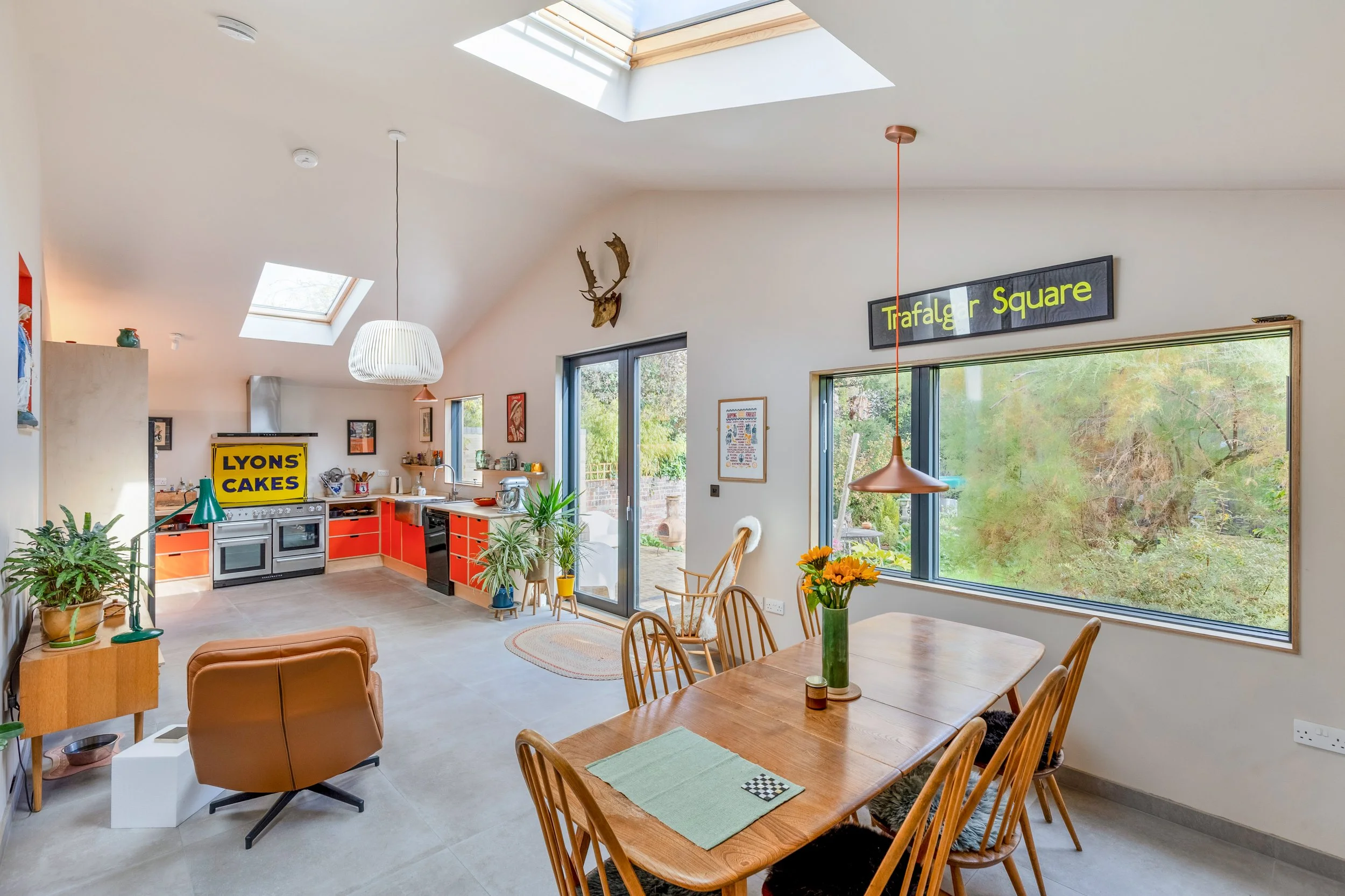 Open-concept kitchen and dining area with skylights and large windows, featuring a wooden dining table with chairs, potted plants, and modern decor.