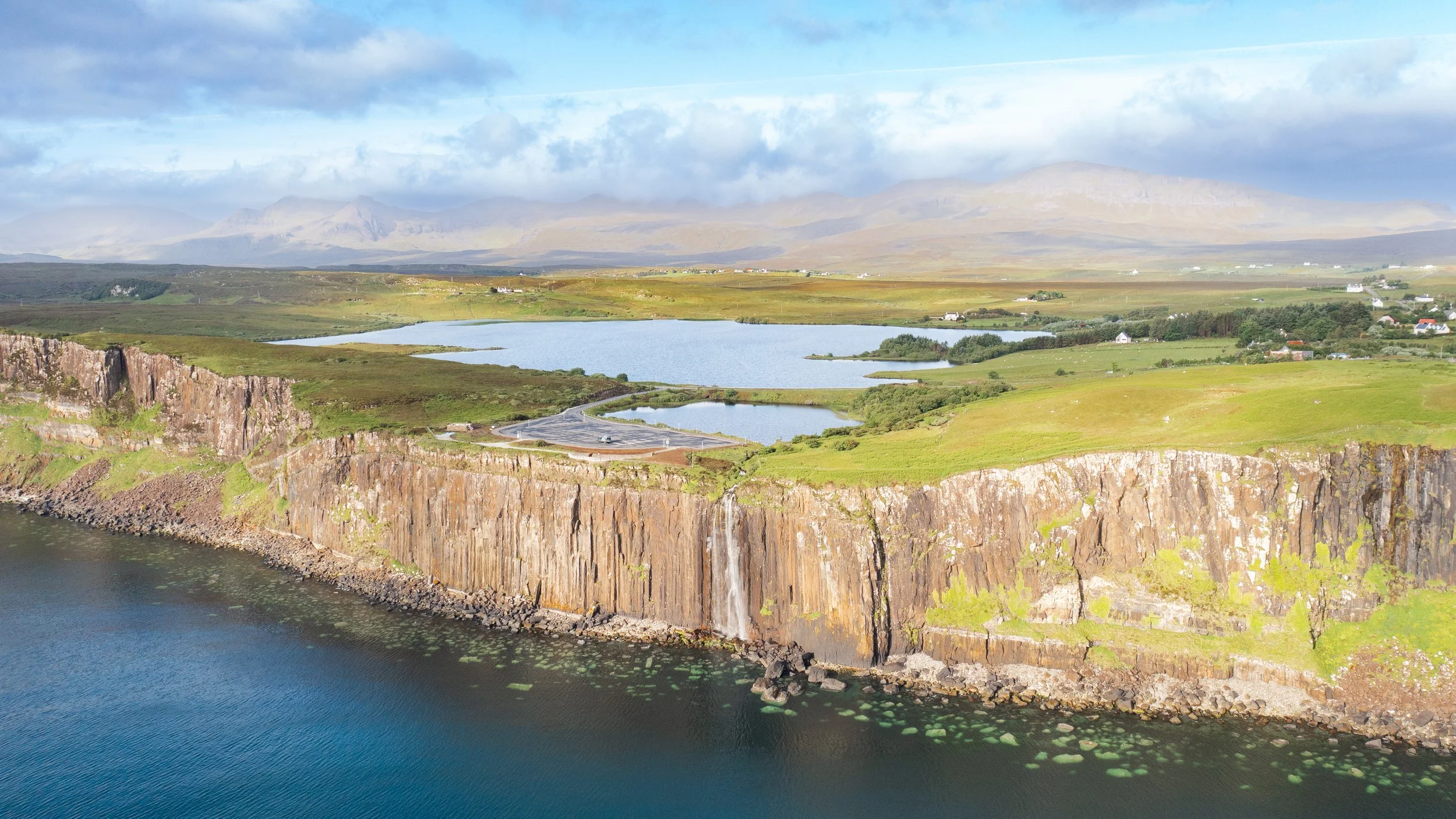A scenic landscape featuring a waterfall flowing from a cliff into a body of water, with rolling green hills, lakes, houses, and mountains in the background under a partly cloudy sky.