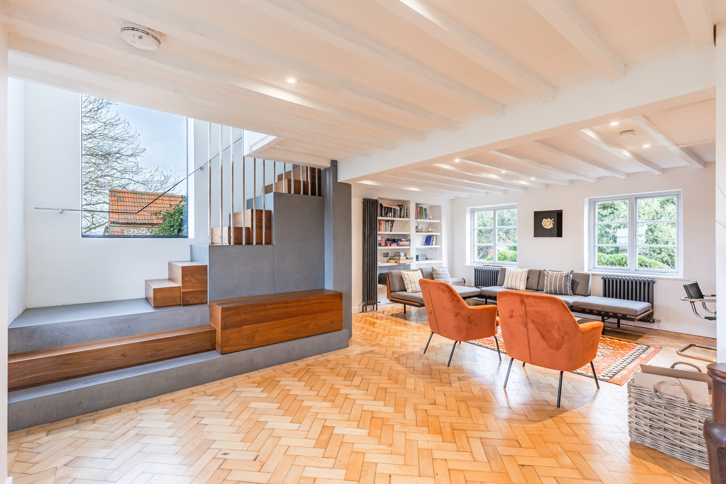 Modern living room with light wood parquet floors, white walls, large windows, and a seating area with gray sofas, orange armchairs, and a bookshelf.