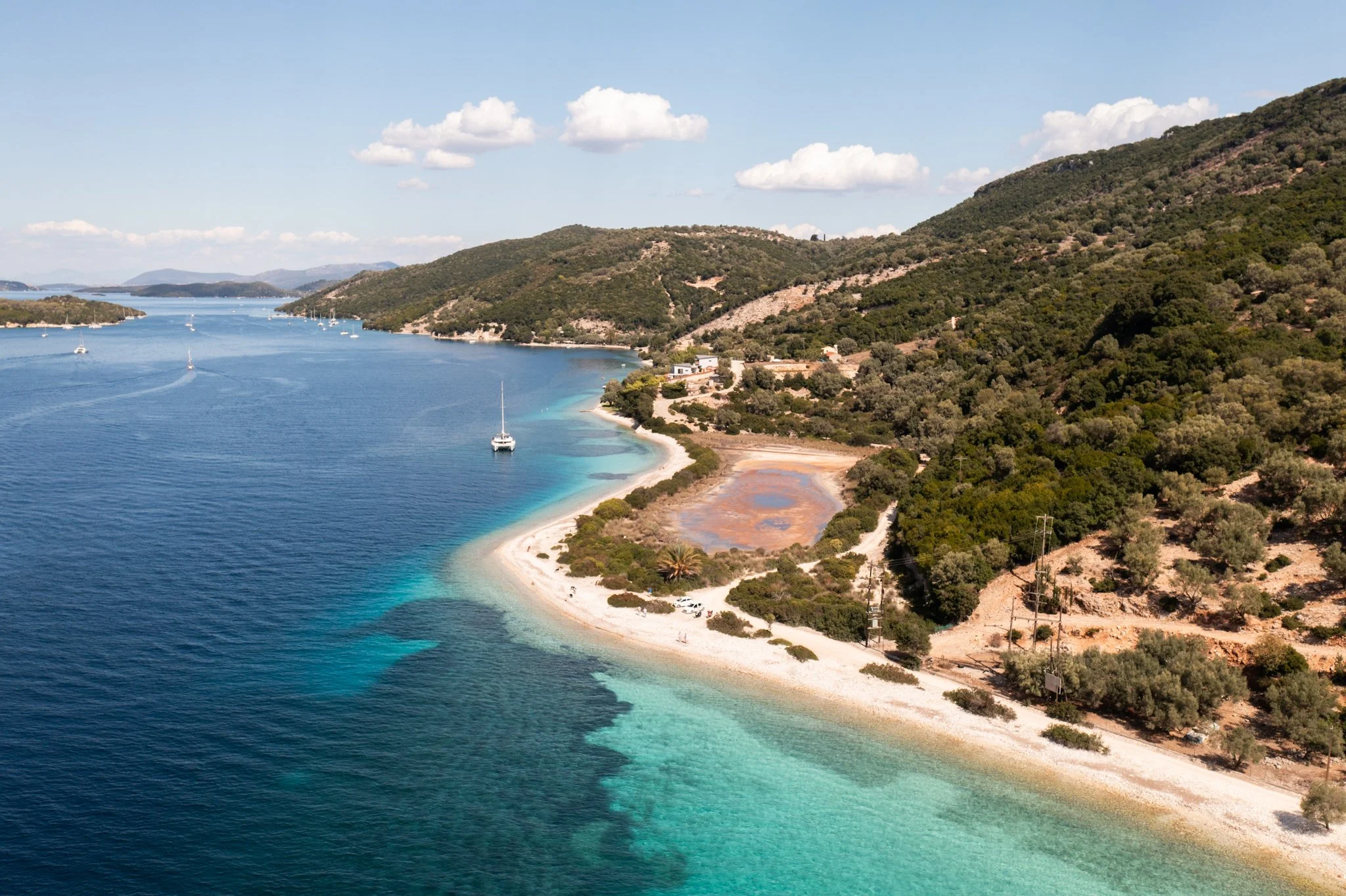 A coastal landscape with a shoreline, clear blue water, sailboats, and green hills in the background under a partly cloudy sky.