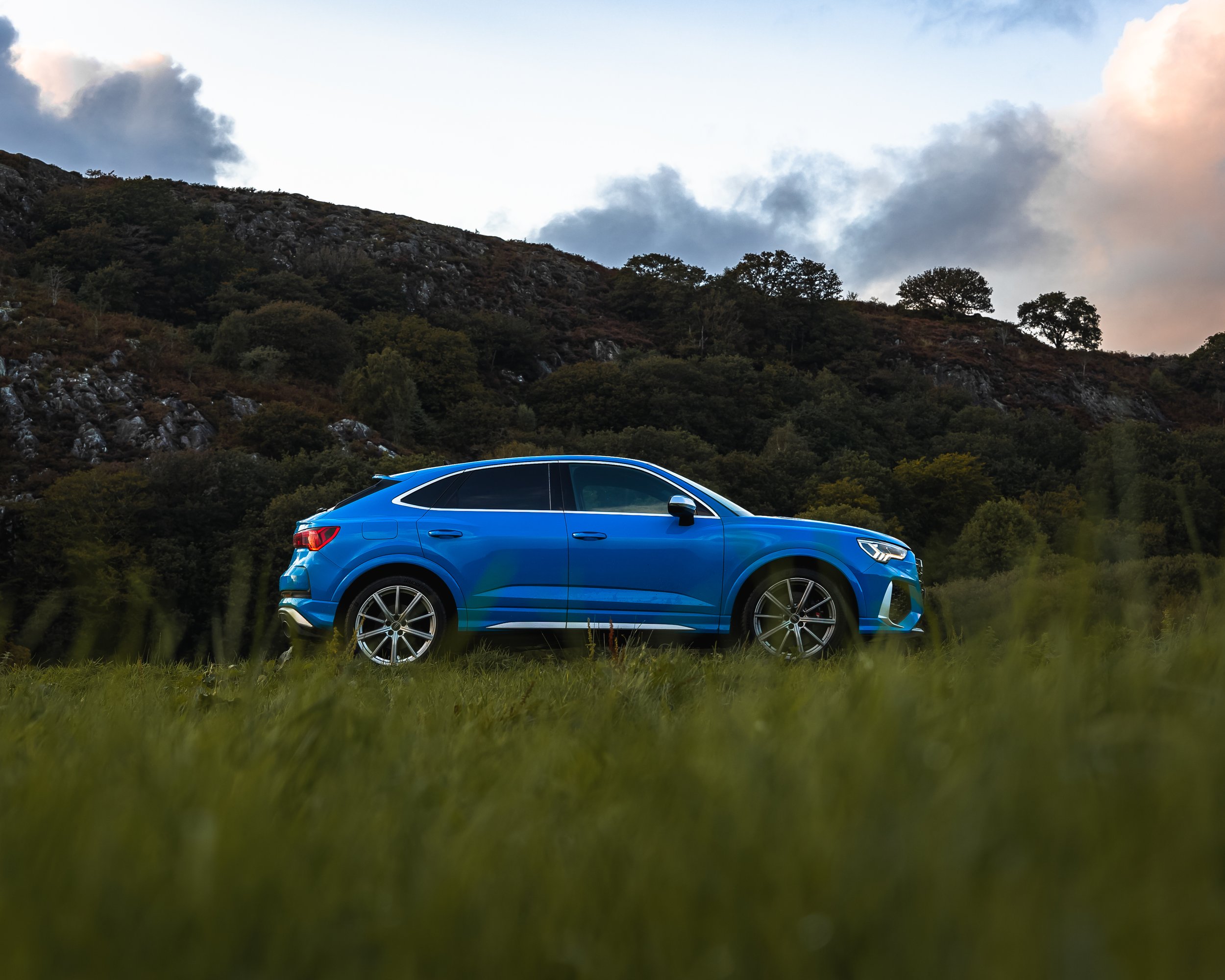 A blue SUV parked on a grassy field with trees and a mountain in the background, under a sky with clouds.