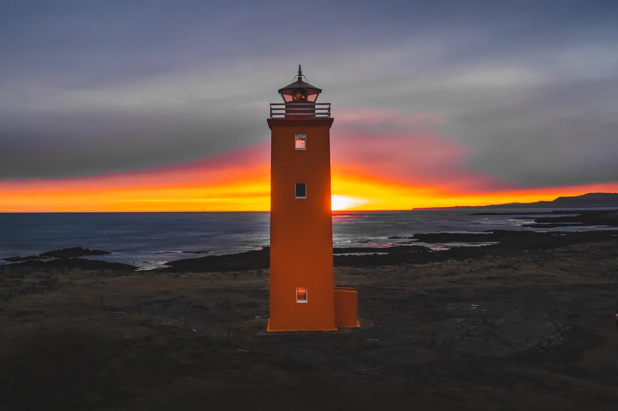 A lighthouse on a coastline at sunset with the sky transitioning from blue to orange and pink colors, and the ocean waves below.