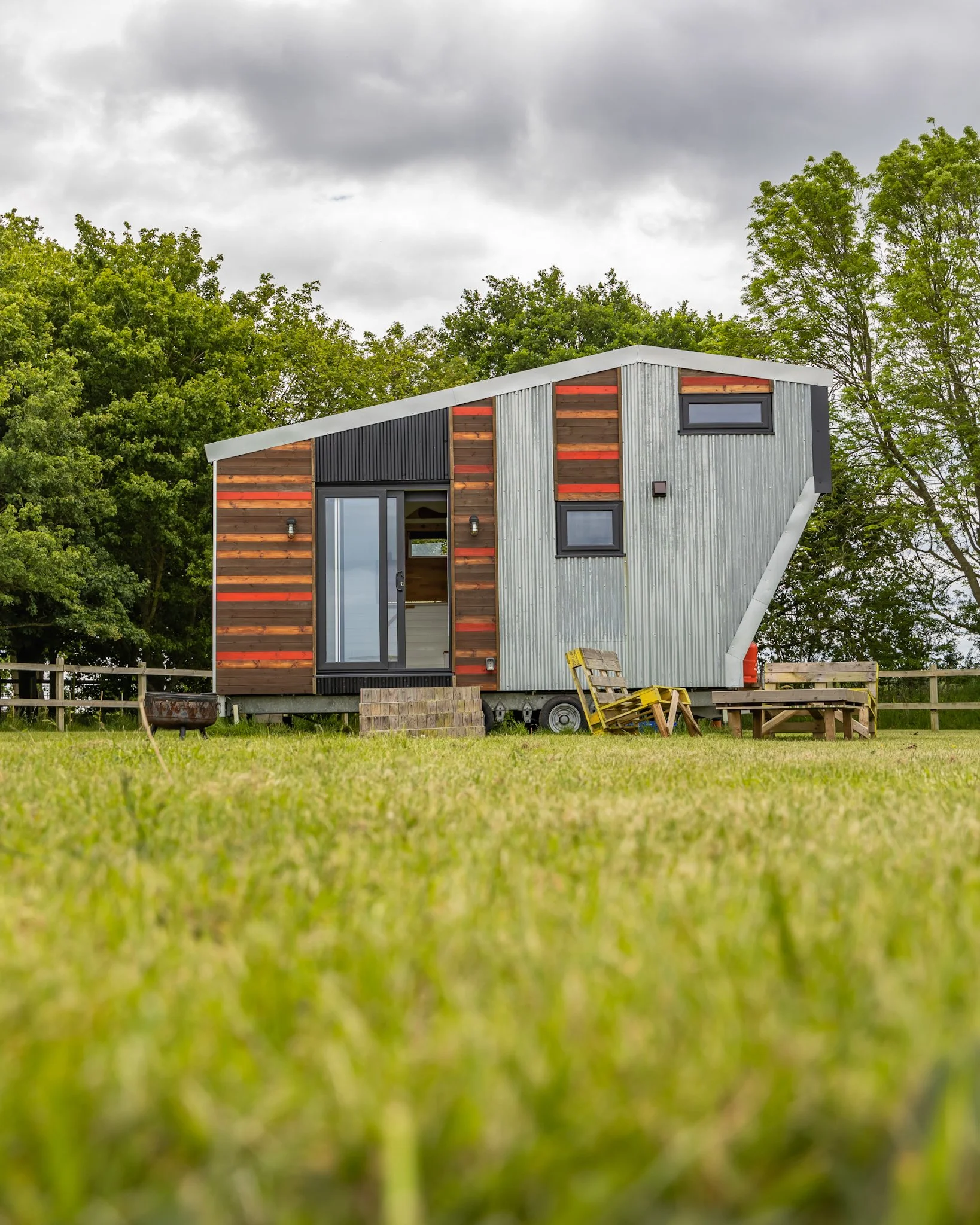 A small, modern house built on a trailer base with a mix of wood and metal exterior, surrounded by green grass and trees, under a cloudy sky.