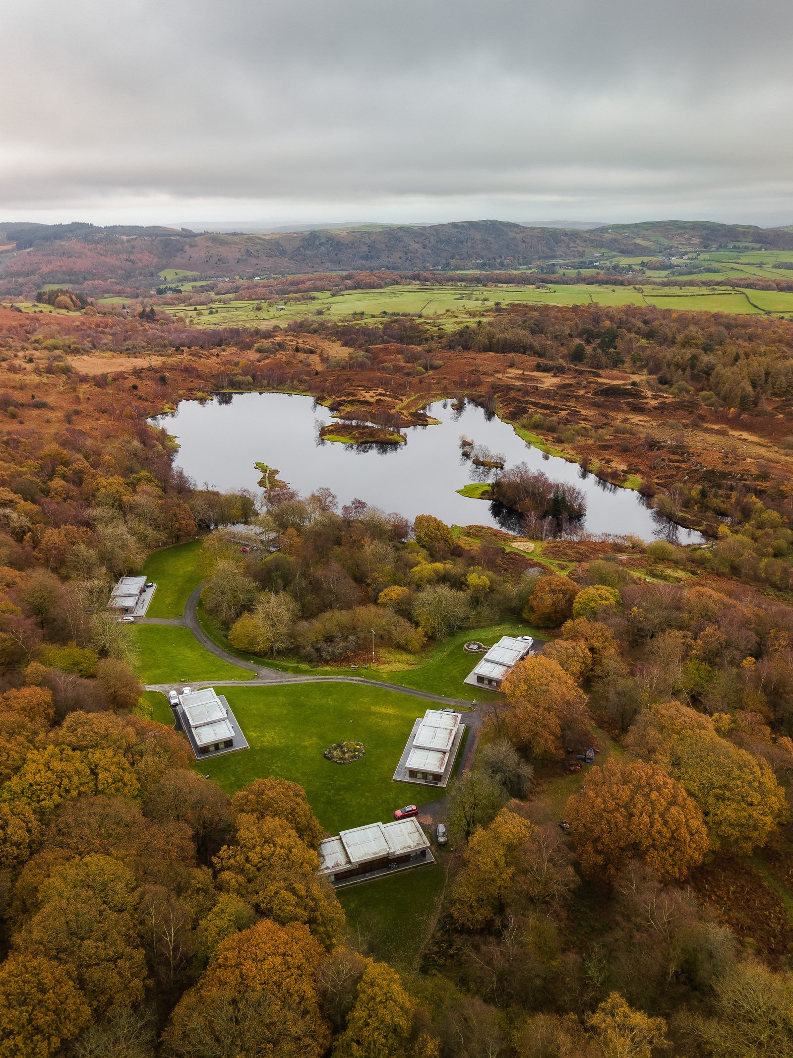 An aerial view of a landscape with a pond, surrounded by autumn-colored trees and modern buildings, with rolling hills and mountains in the background under a cloudy sky.