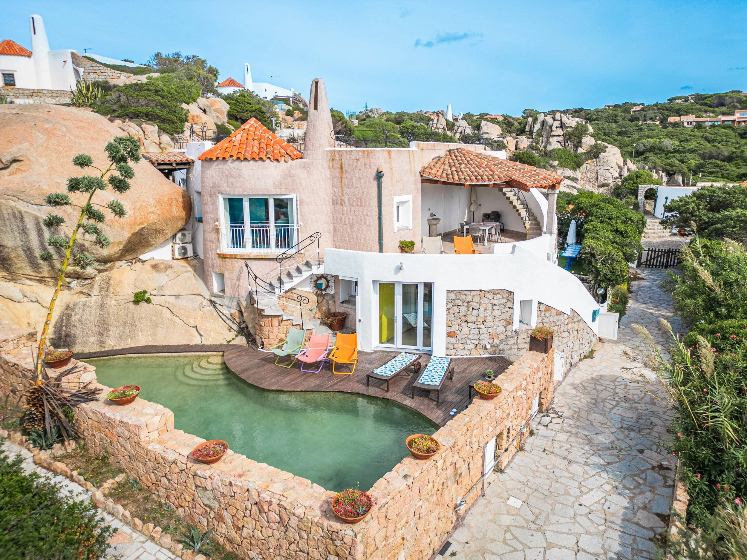 A hillside Villa with a white exterior and orange tiled roof, a small outdoor patio with chairs, and a curved swimming pool outlined by a stone wall with potted plants.