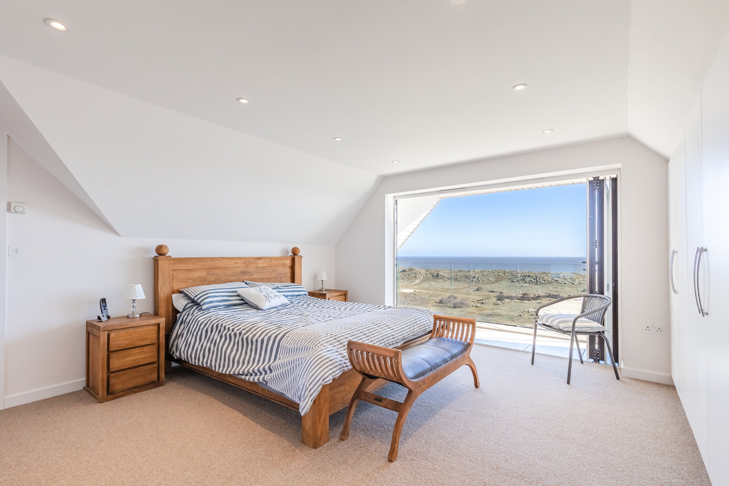A bedroom with a large window overlooking a coastal landscape, a wooden bed with striped bedding, bedside tables with lamps, and a chair.