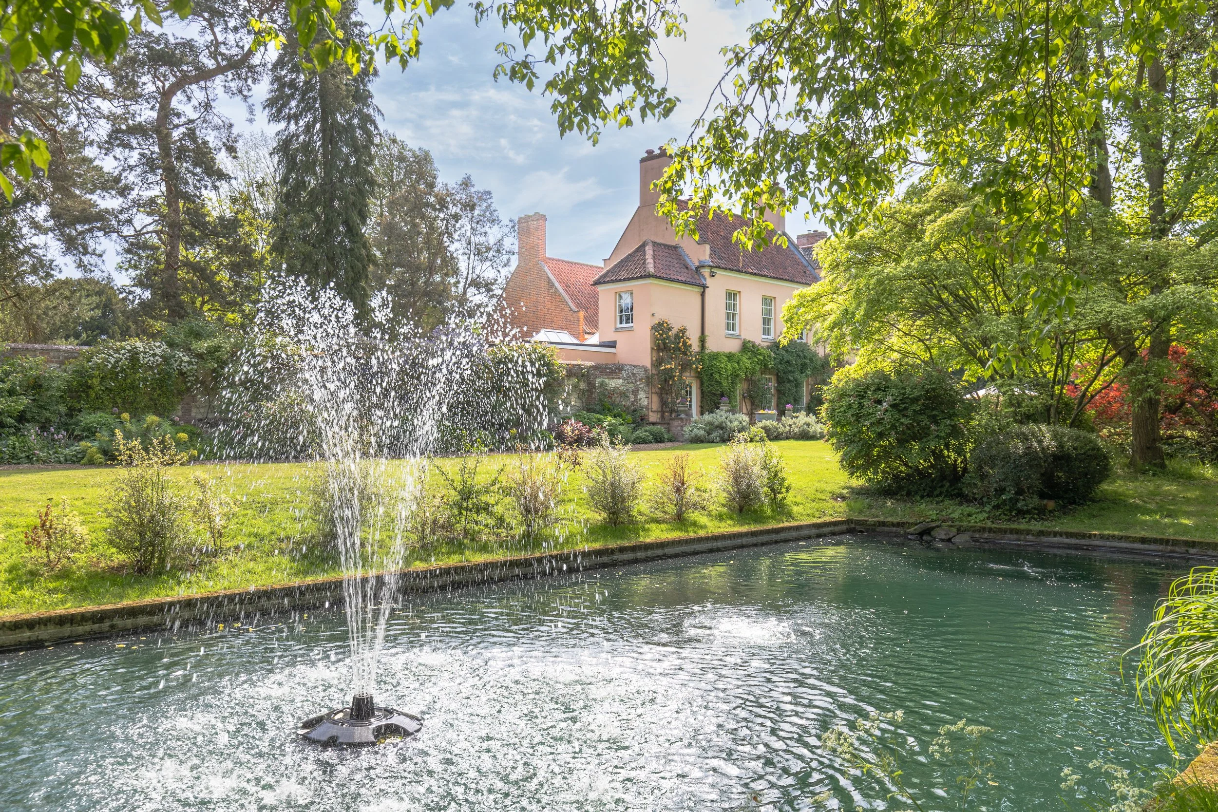 A backyard scene with a pond and a fountain, surrounded by green trees and shrubs. In the background, there are houses with red tile roofs and a chimney, under a partly cloudy sky.