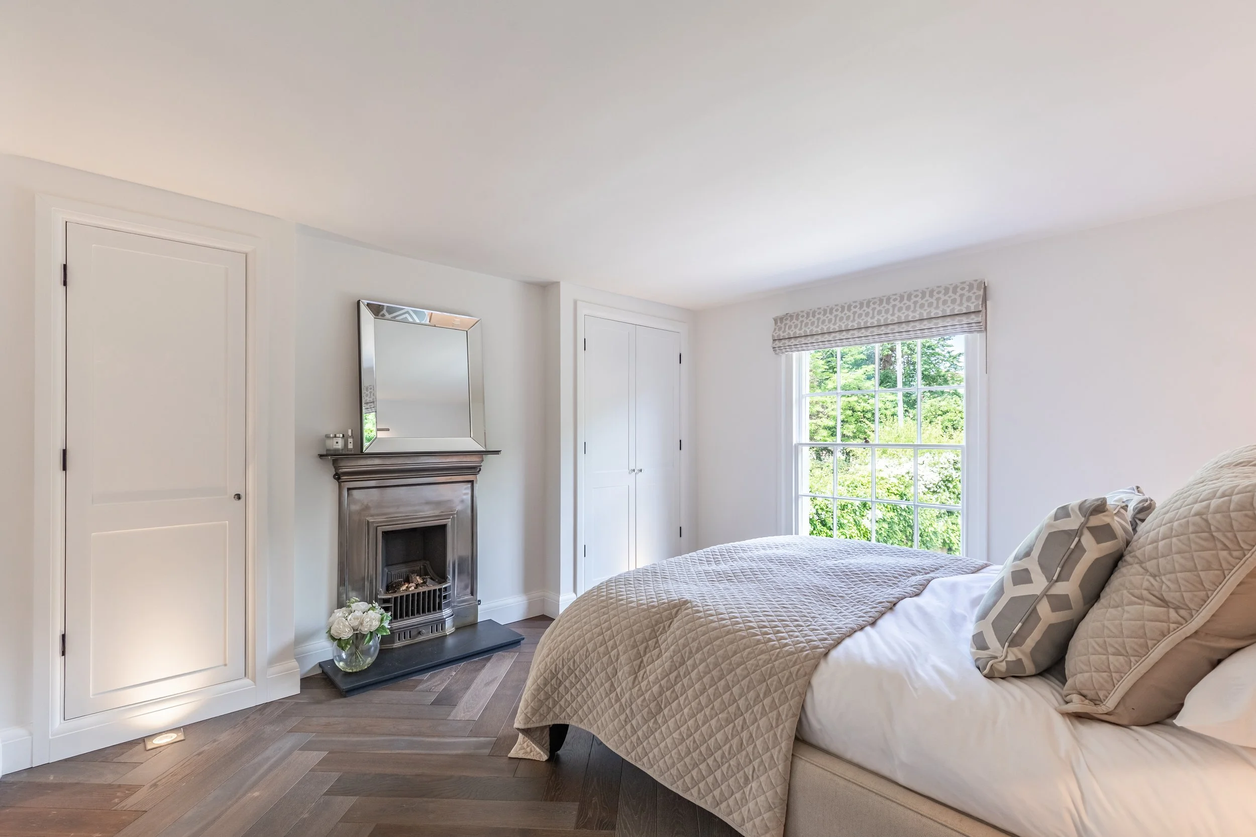 A cozy bedroom with a large window showing green trees outside. The bed has a beige quilted coverlet and patterned pillows. There is a small fireplace with a mirror above it, a door on the left, and a white double closet to the right of the fireplace