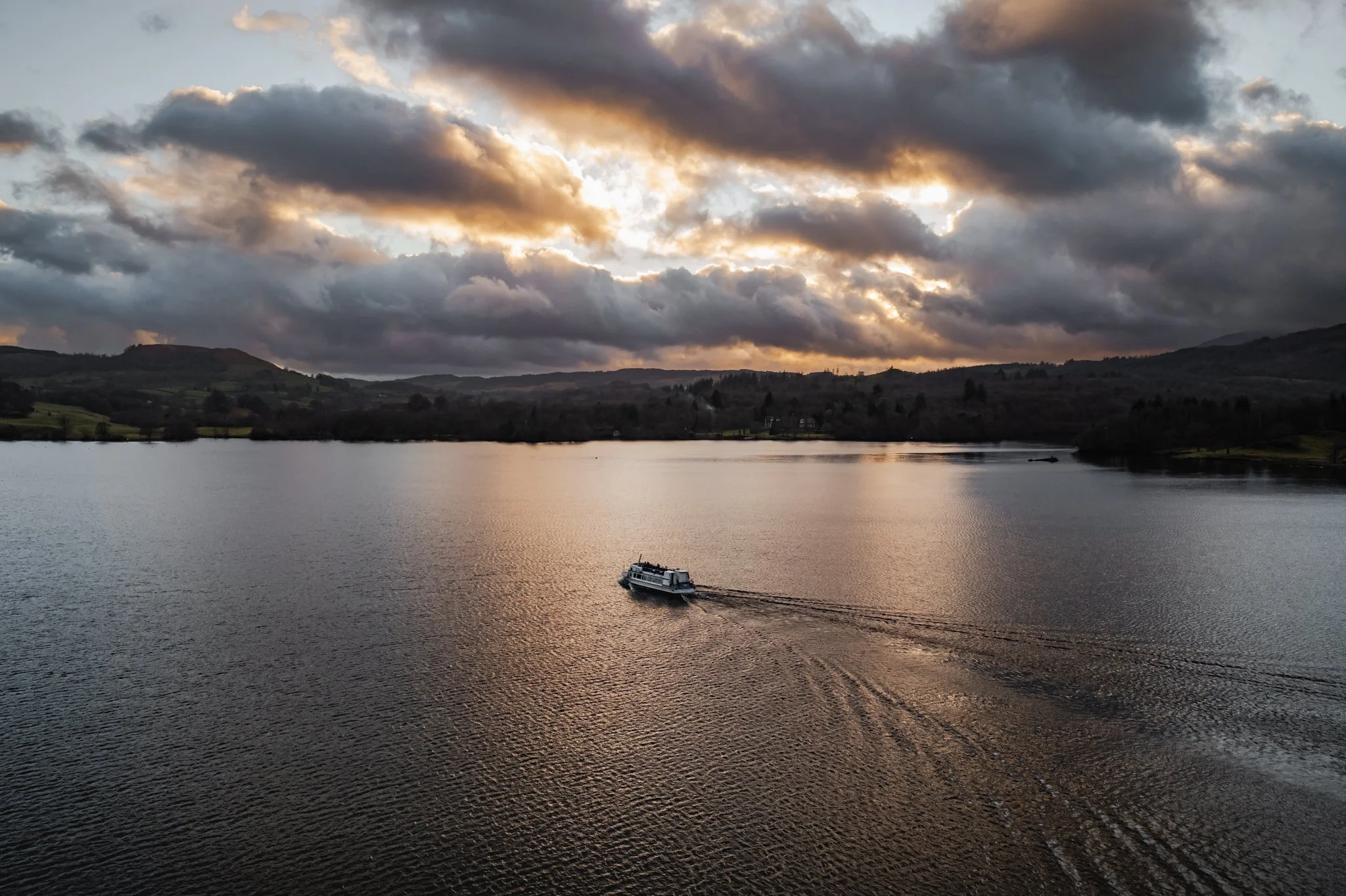 A boat moving across a calm body of water during a cloudy sunset, with rolling hills in the background.