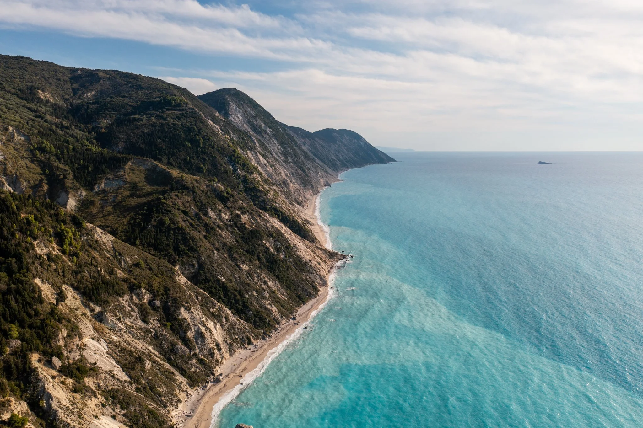 Coastal cliffs with green vegetation rising from turquoise sea, and a small island in the distance under partly cloudy sky.