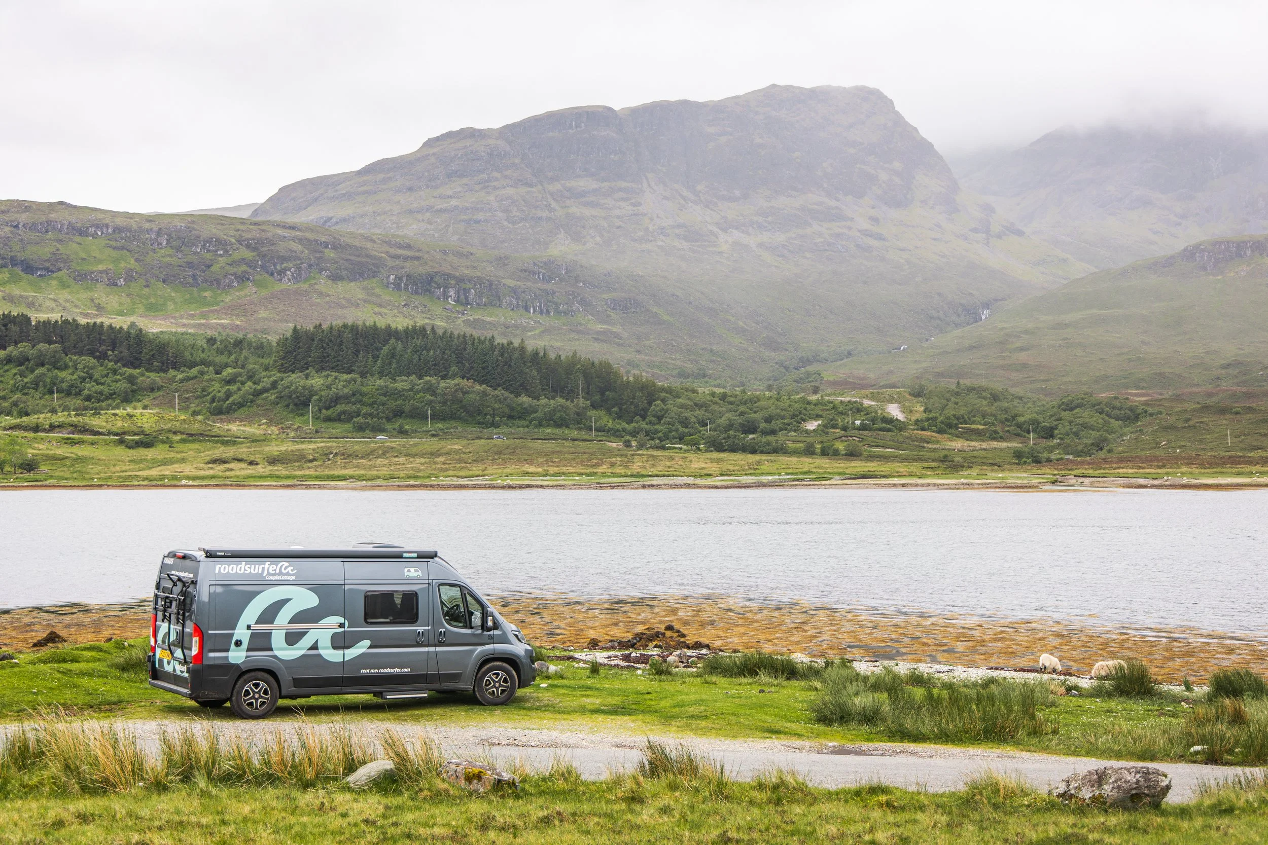 A scenic landscape featuring a mountain range in the background, a body of water, and a green grassy area with a gray camper van parked near the water.