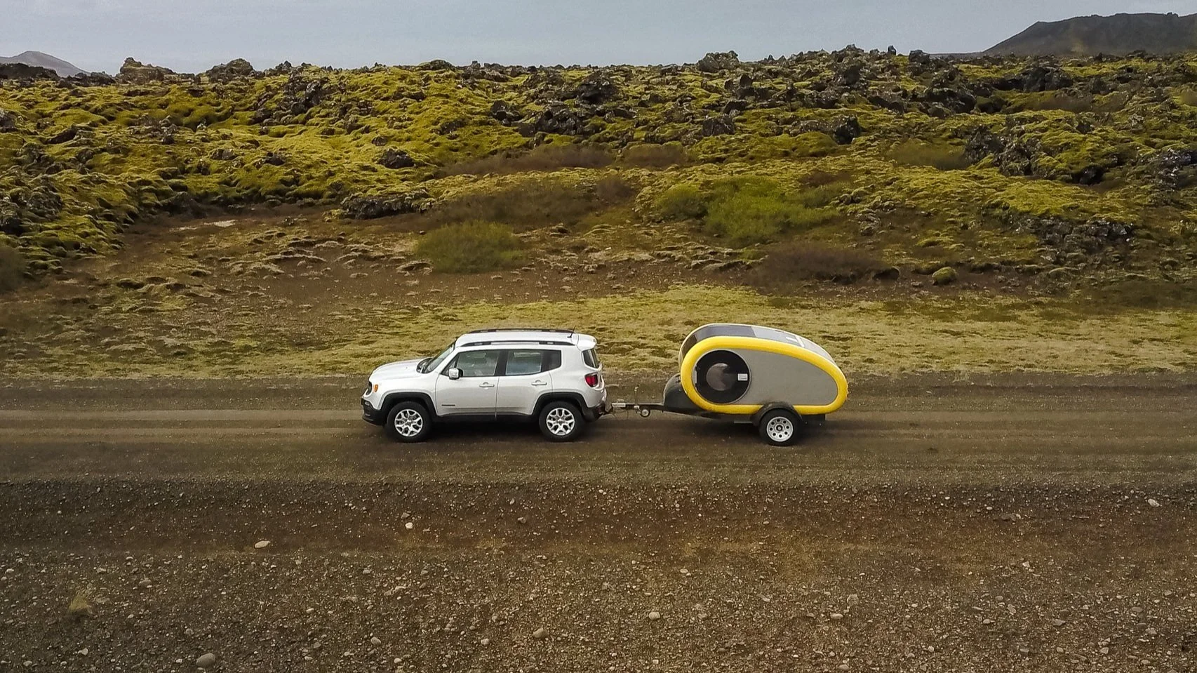 A white SUV towing a small portable camper trailer on a dirt road with a rugged moss-covered landscape in the background.