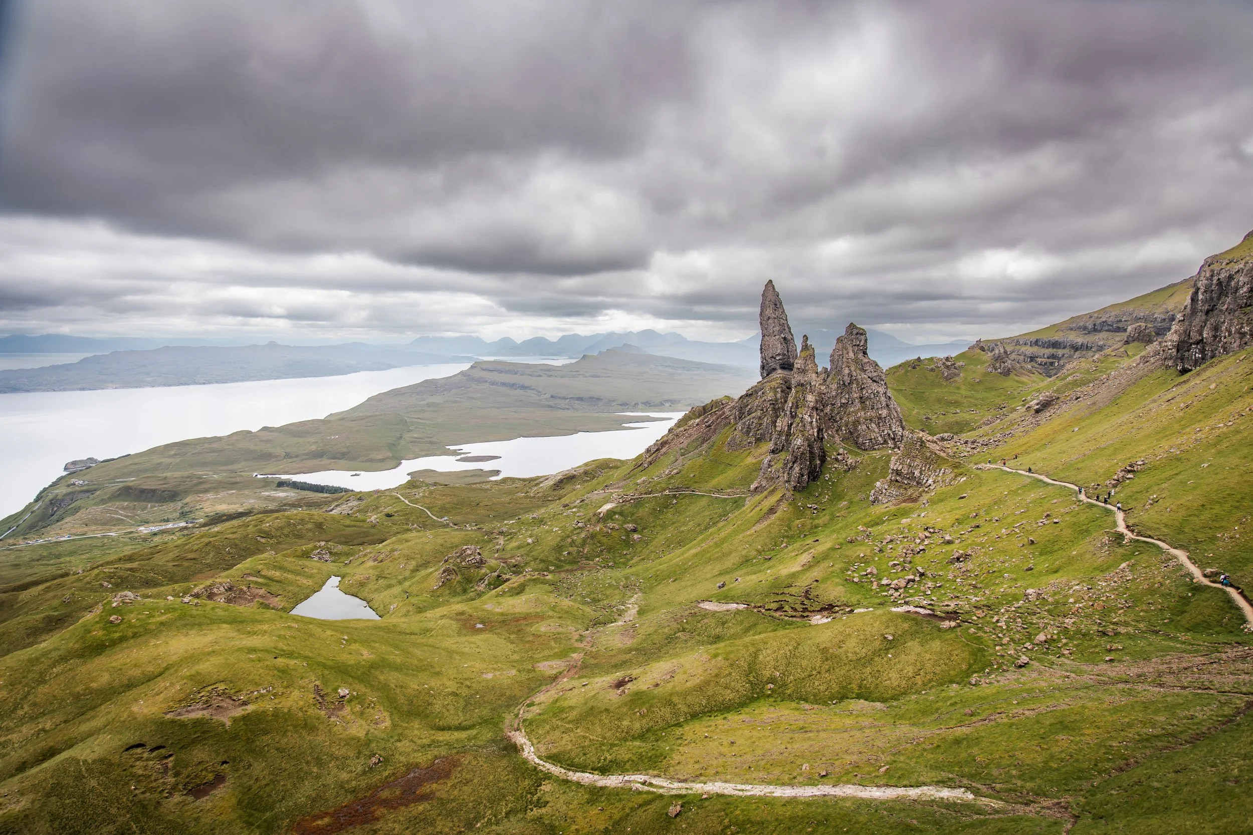 A scenic landscape featuring green rolling hills, a large body of water, and dramatic cloudy skies with a tall, narrow rock formation in the foreground.