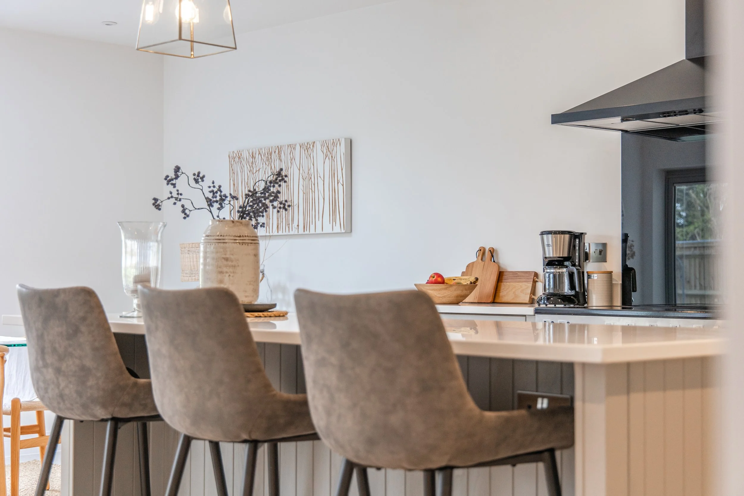 Modern kitchen with a white countertop, three upholstered chairs, a decorative vase with branches, a wooden tray with fruit, and kitchen appliances including a coffee maker and a wall-mounted microwave. The background has a white wall with a piece of