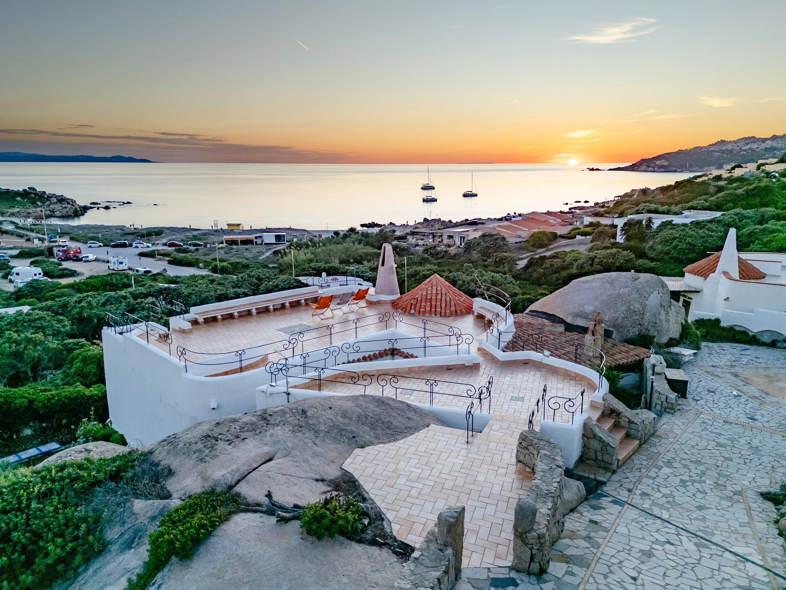Sunset view over a coastal town with sailboats on calm water, terracotta-roofed buildings, and greenery.