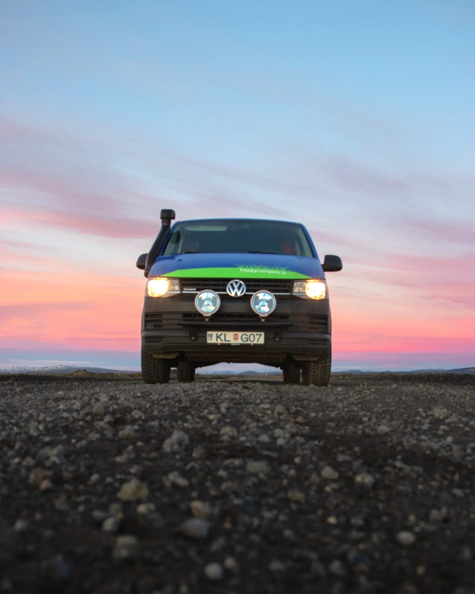 Front view of a black Volkswagen van parked on a gravel road during sunset, with a colorful sky in the background.
