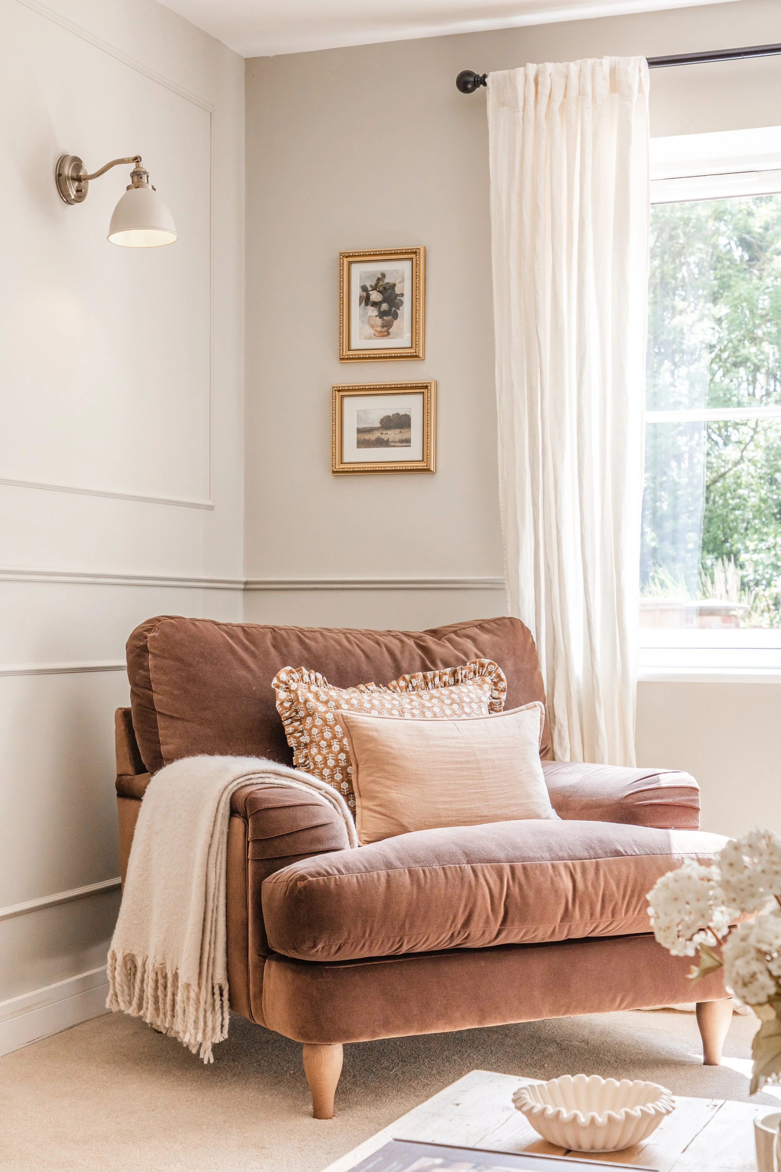 Living room with a dusty pink armchair with four pillows, a beige blanket draped over the armrest, white curtain, two framed pictures of nature, and a window with green trees outside.