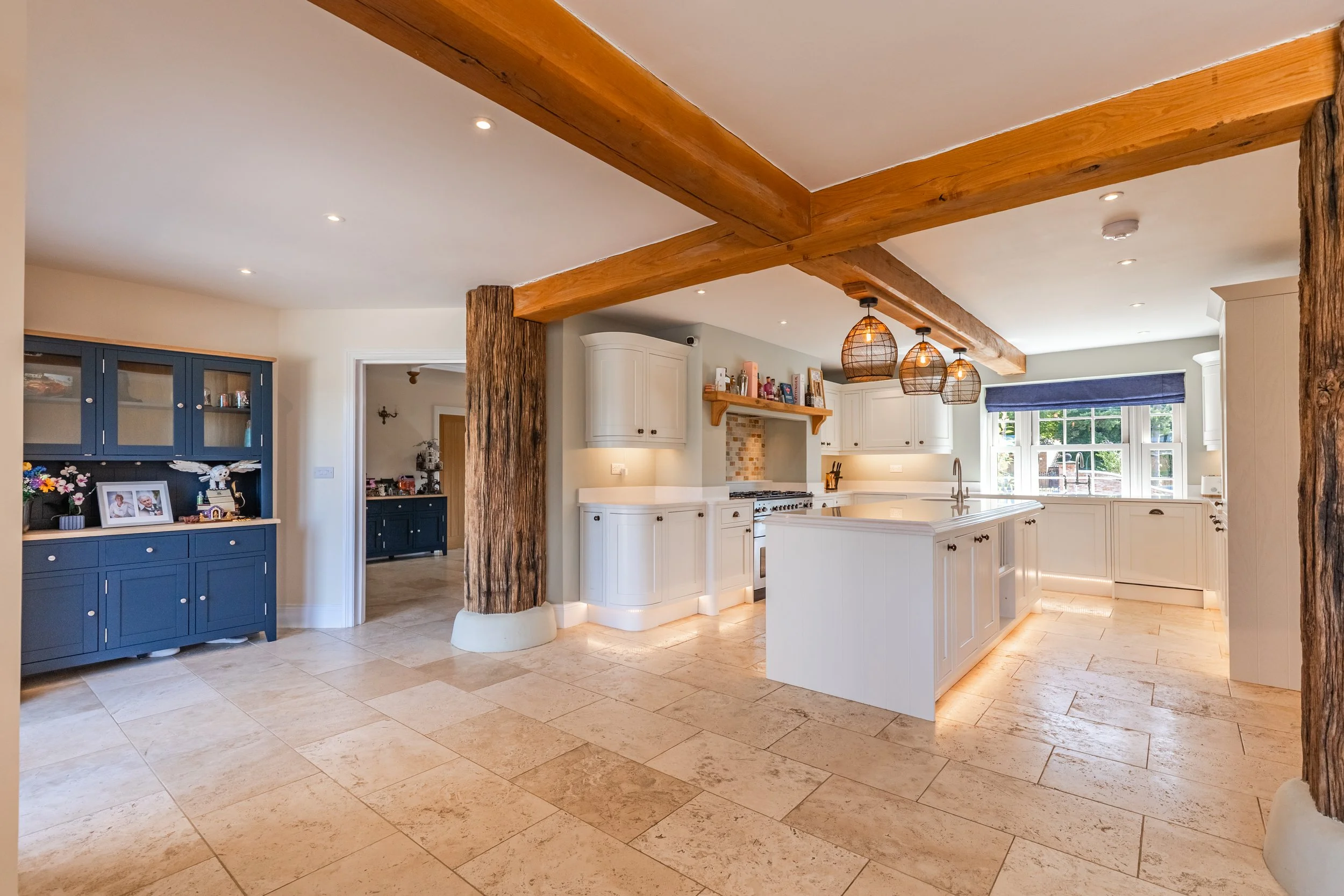 A spacious, open-plan kitchen with cream-colored cabinetry, a large kitchen island, and natural wood beams on the ceiling supported by rustic wooden poles. The kitchen features pendant lights and large windows with a view of greenery outside.