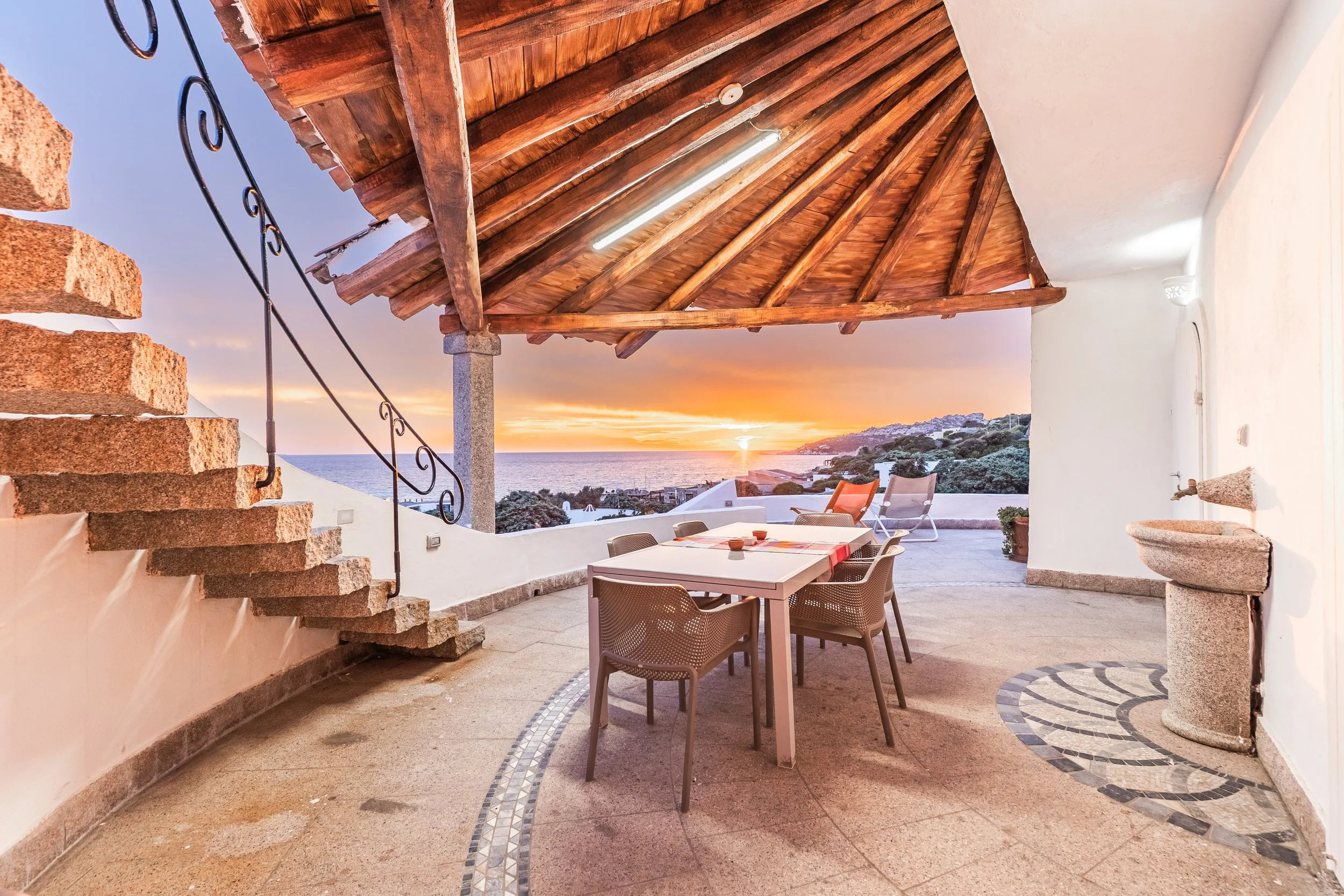 An outdoor patio area with a table and chairs overlooking the ocean at sunset. The patio has a wooden roof, stone stairs, a decorative stone basin, and walls with white paint.