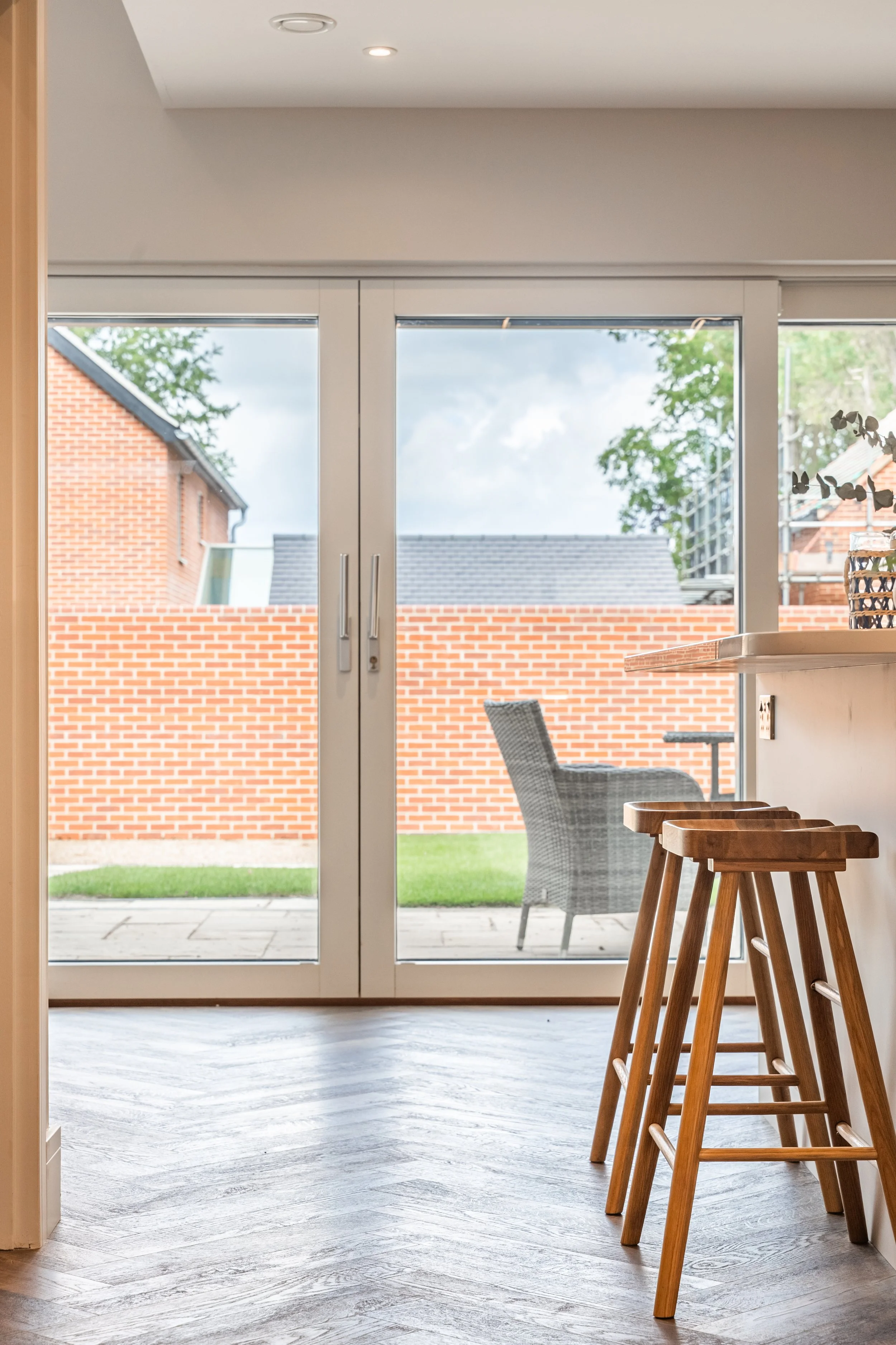 View of a modern kitchen with dining area, highlighting four wooden bar stools, a sliding glass door leading to an outdoor patio, and a brick wall outside.