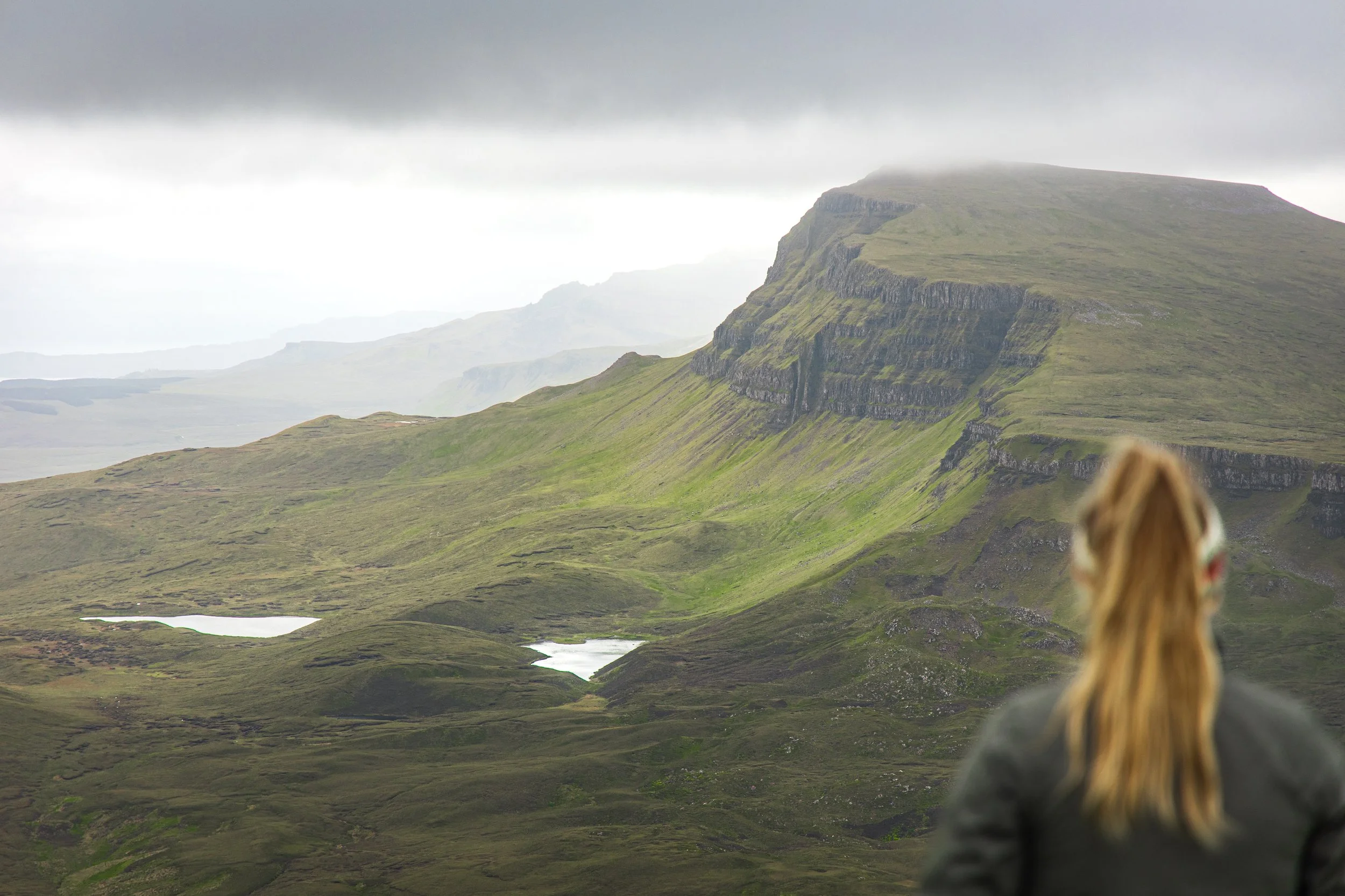 A person with long hair, wearing a gray jacket, looking at a mountainous landscape with green hills, rocky cliffs, small lakes, and overcast skies.