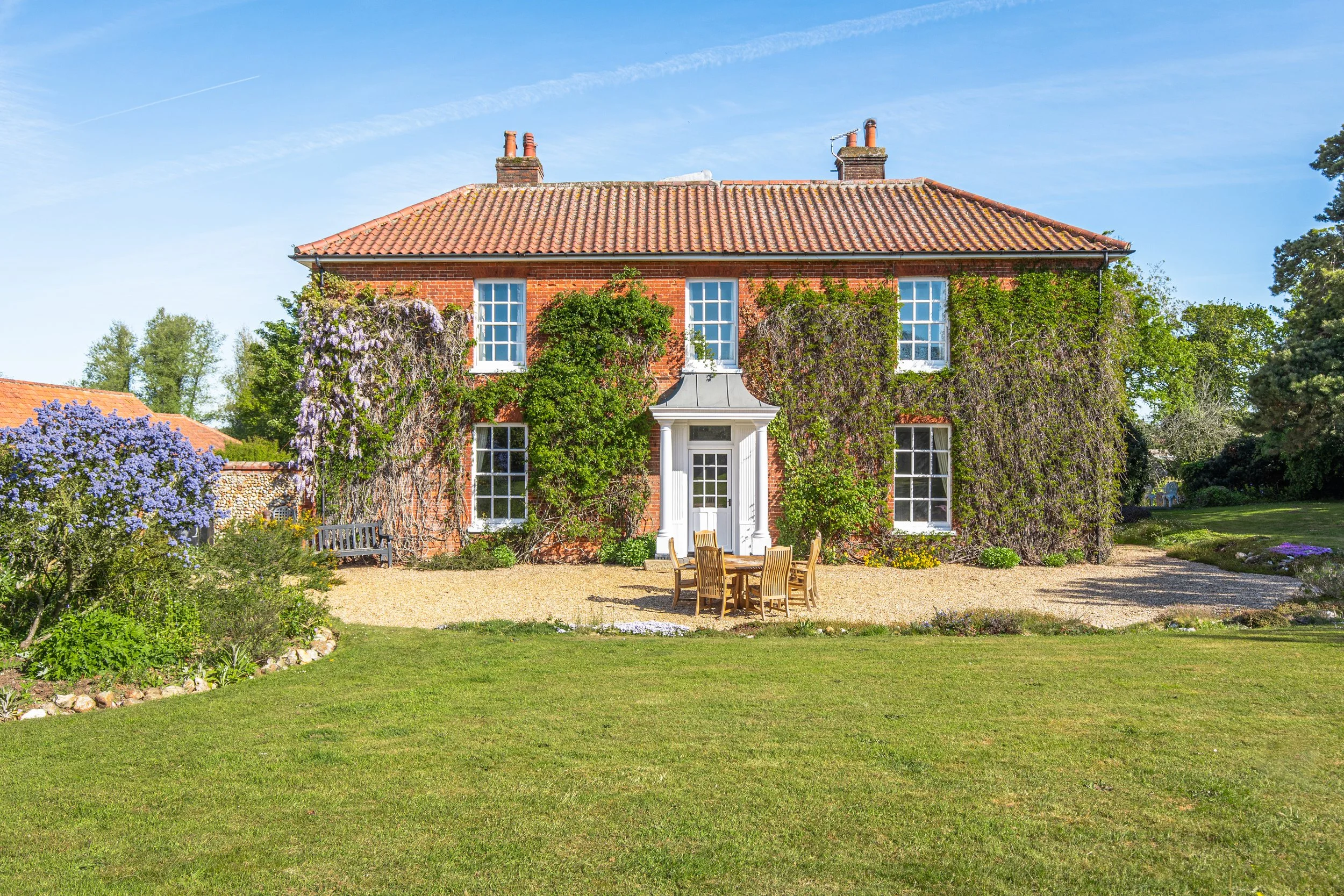 A large brick house with a red tile roof, surrounded by green trees and gardens, with a patio table and chairs in the yard under a blue sky.