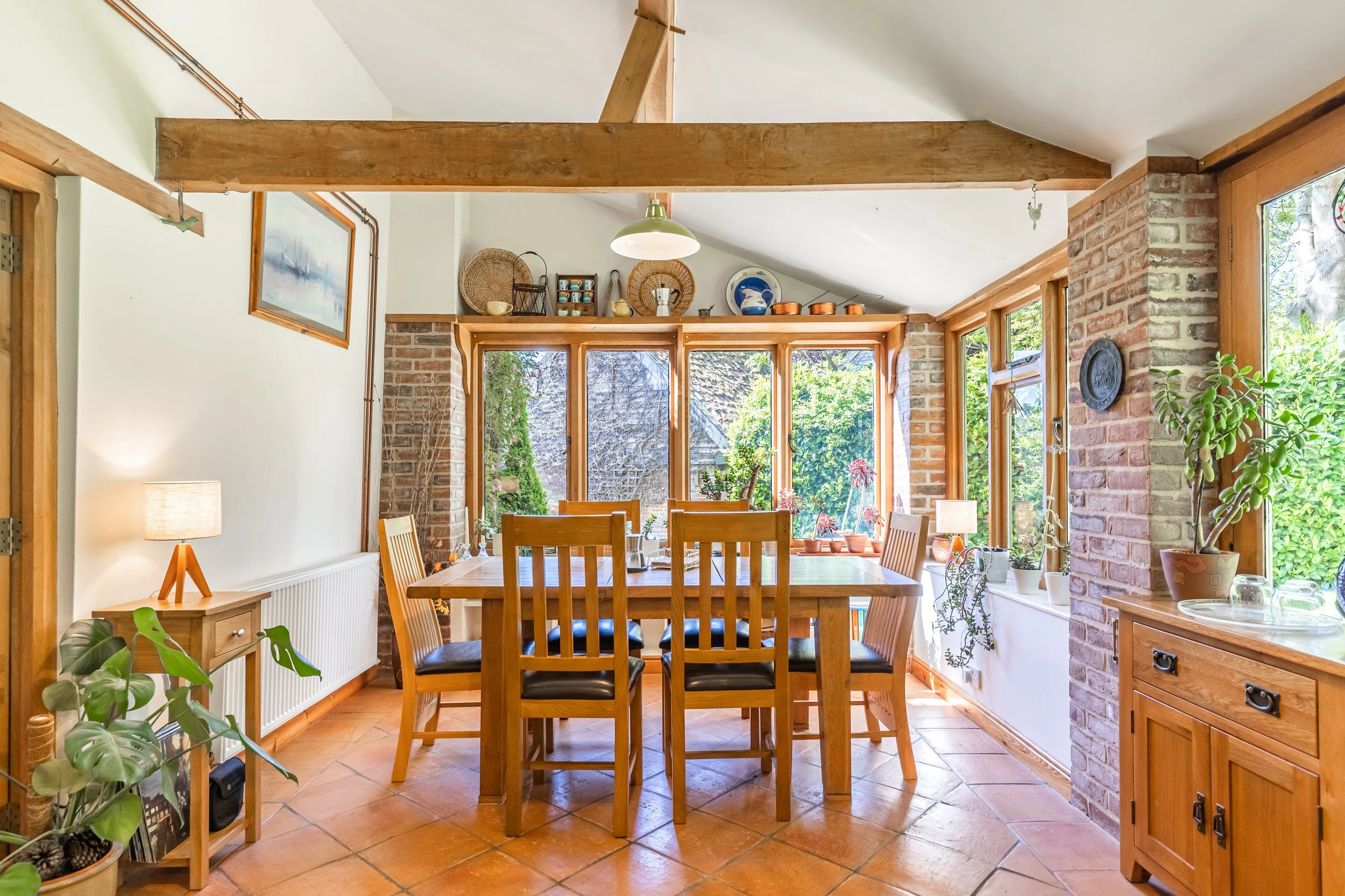 The image depicts a cozy dining room with a wooden table and six matching chairs. Large windows let in natural light, revealing a garden outside. The room has exposed brick walls, wooden trim, and various decorative items on a high shelf, including b