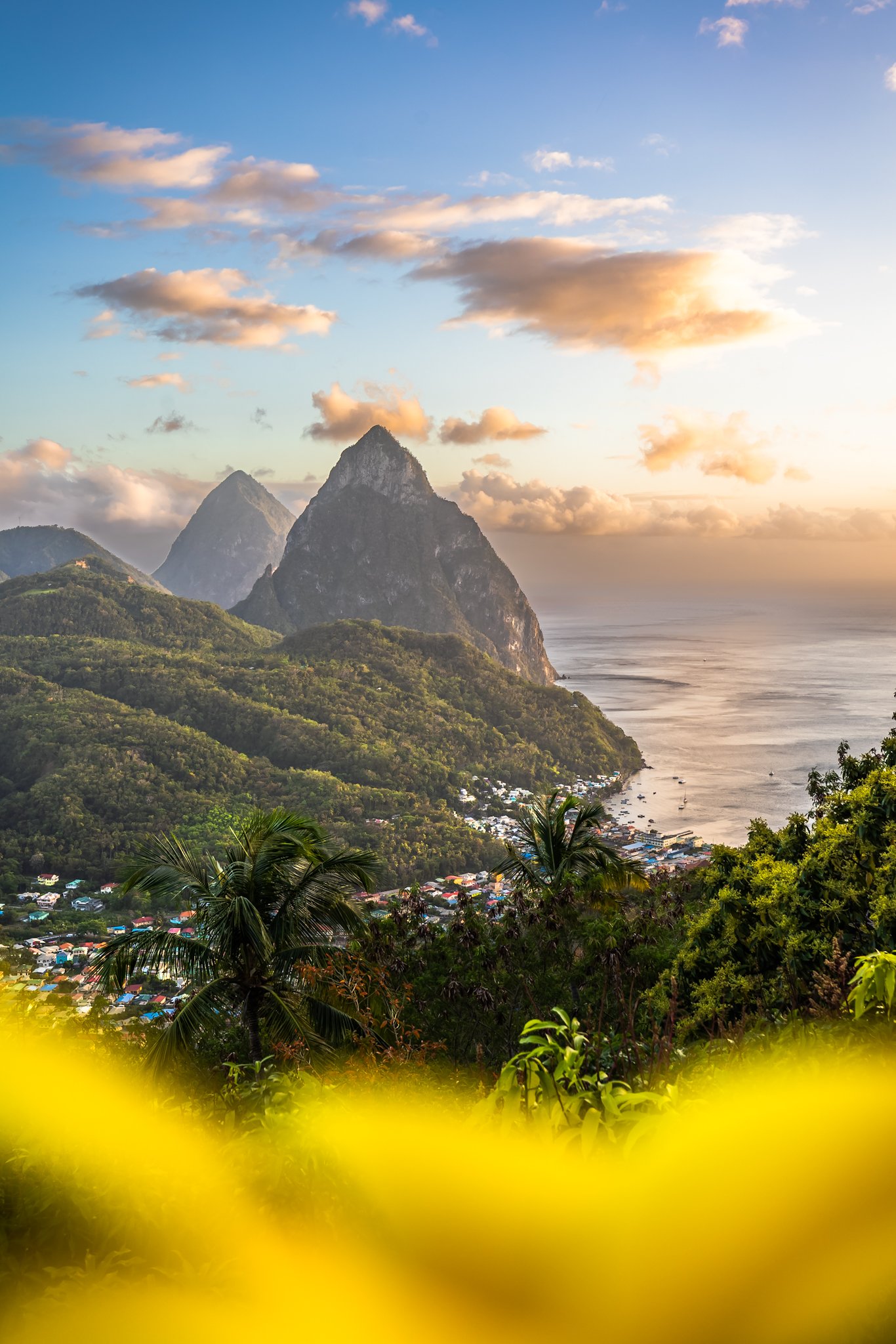 Scenic view of lush green mountains and a coastal village with boats, overlooking the sea during sunset, with a cloudy sky overhead.