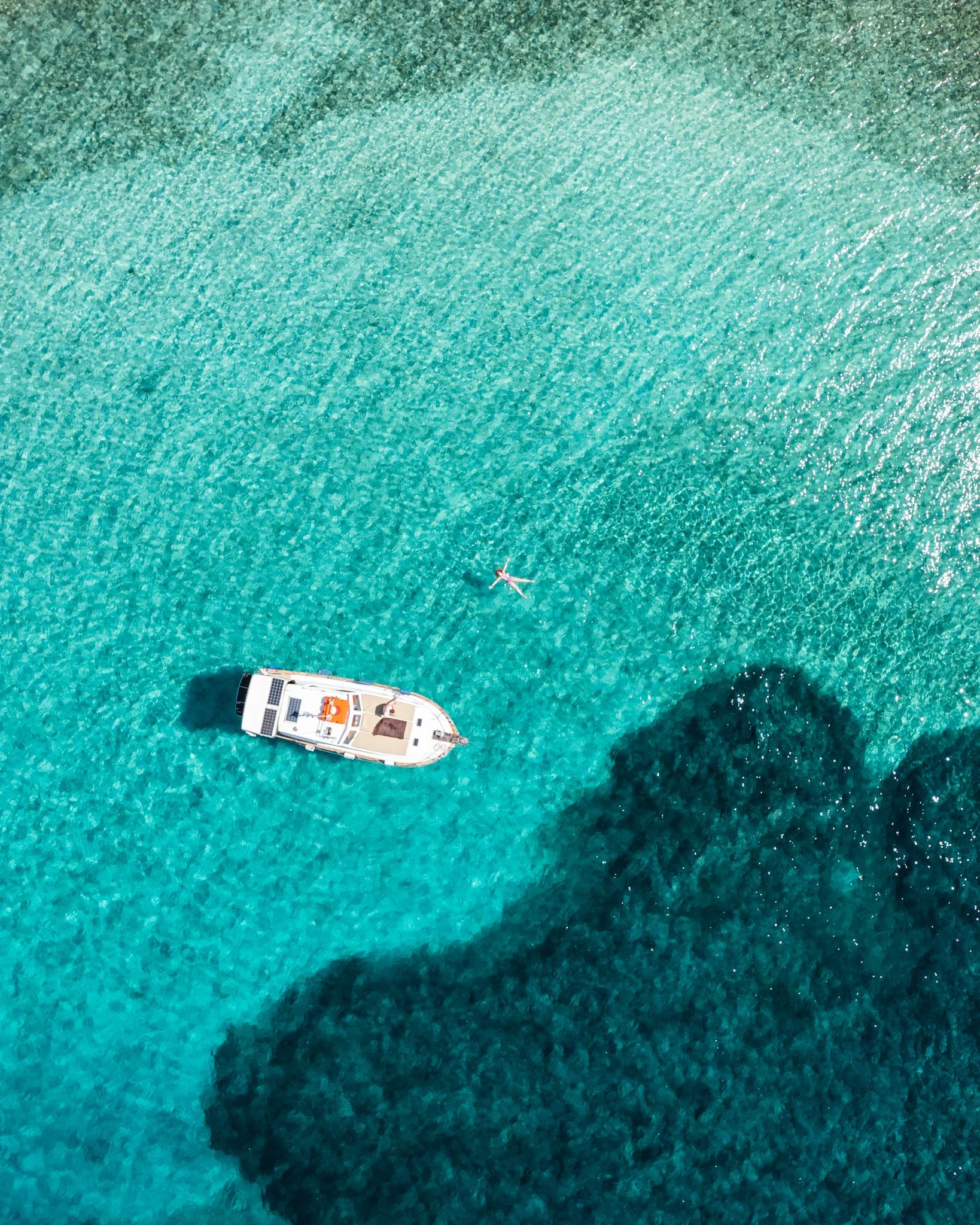 An aerial view of a person floating on their back in clear blue water near a yacht, with shadows from the yacht cast on the water.