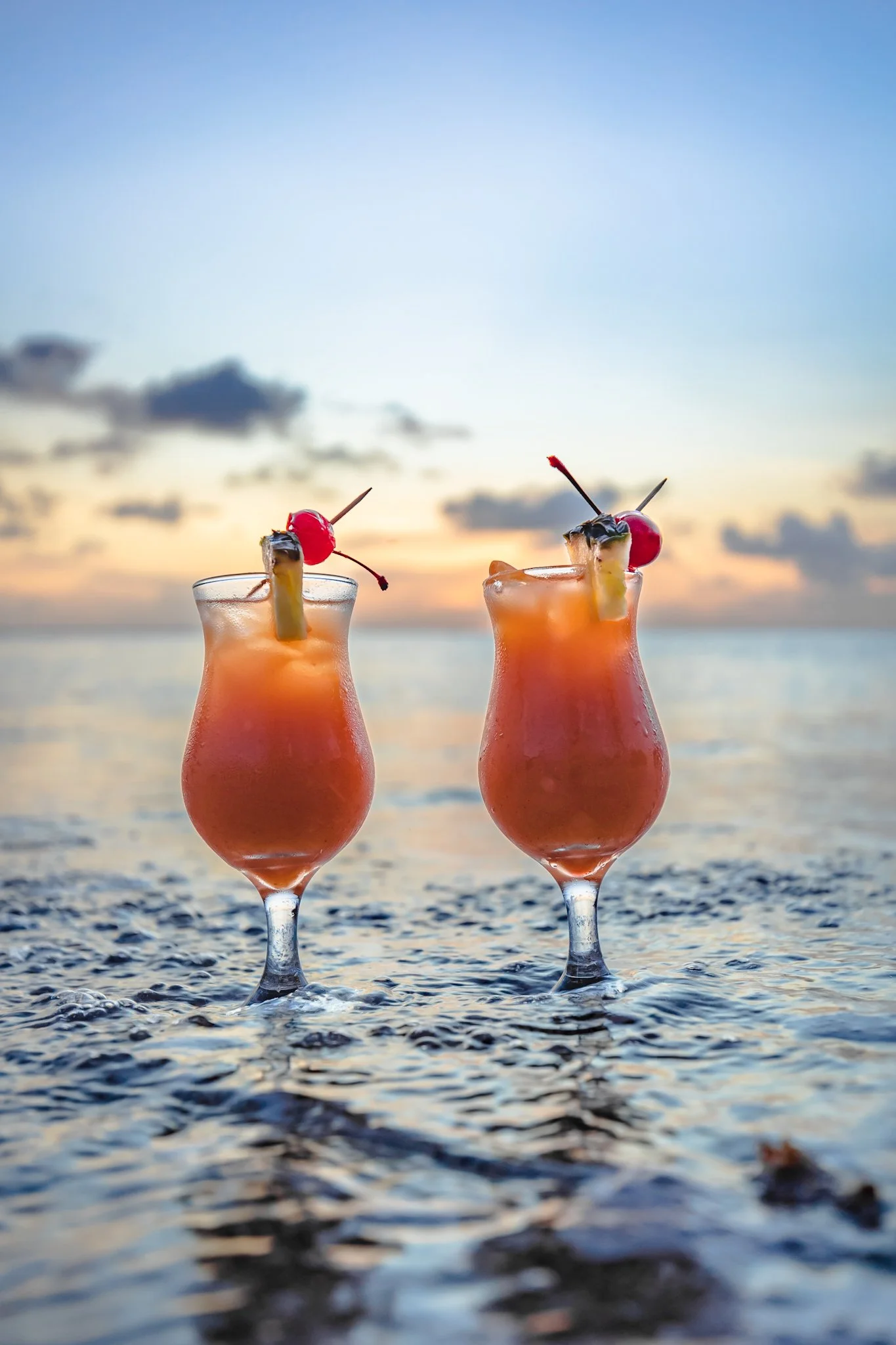 Two pink tropical cocktails with cherries and pineapple garnishes sit on the edge of the water at sunset, with clouds in the sky.