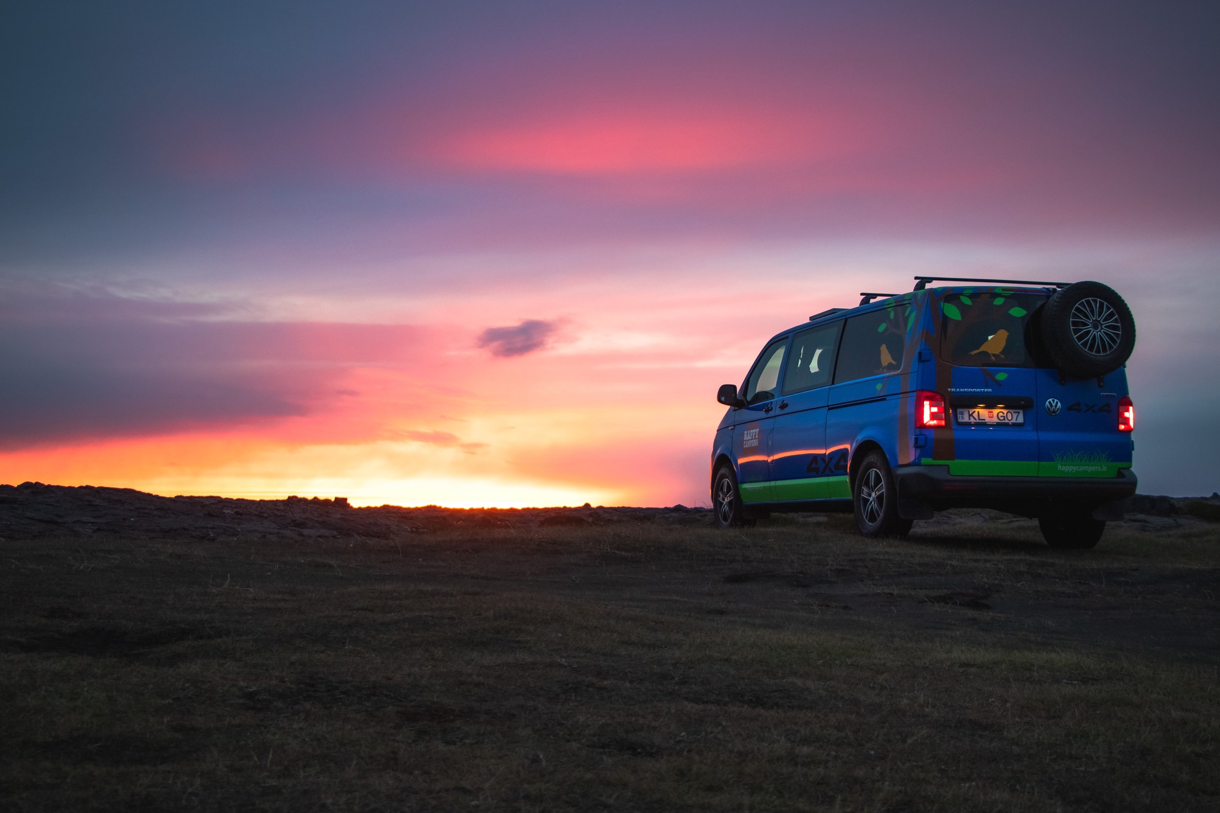 A blue Volkswagen camper van with colorful graphics and a spare tire on the back, parked on a rocky terrain during sunset with a colorful sky and clouds.